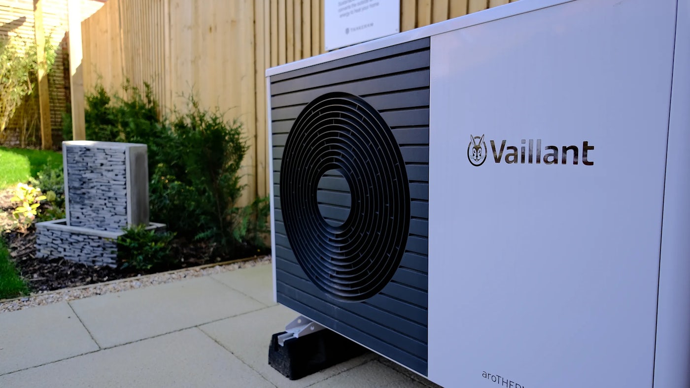A white Vaillant aroTHERM air-to-water heat pump with a black circular vent on the front, placed outdoors on a concrete surface. The Vaillant logo is visible on the side. Behind the unit, a wooden fence and some greenery are present.
