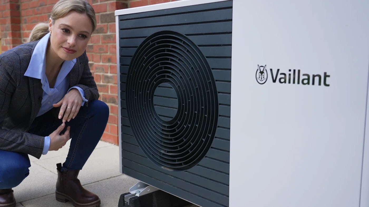 A person crouches next to a Vaillant heat pump unit installed outdoors against a brick wall. The unit has a circular vent with concentric rings on the front, and the Vaillant logo is visible on the right side. The setting suggests a focus on modern heating solutions, energy efficiency, and environmental sustainability.