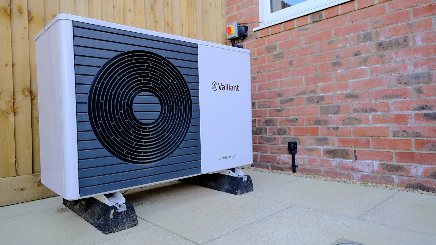 A Vaillant aroTHERM plus outdoor heat pump unit installed next to a brick wall and a wooden fence. The unit has a circular vent design on the front and is elevated on concrete blocks. An electrical switch is mounted on the wall above it.