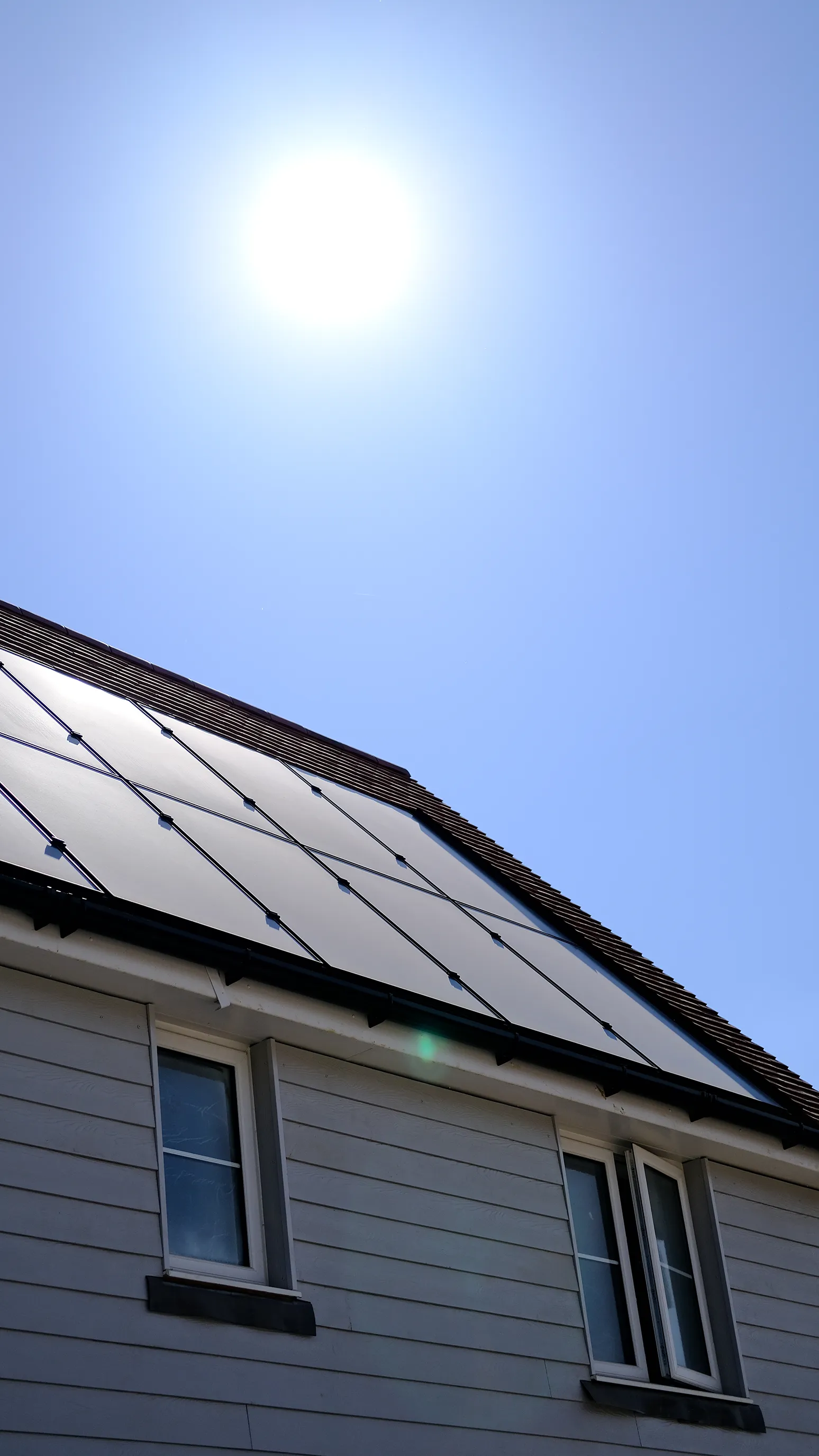 A house with solar panels installed on its roof under a bright, clear sky. The sun shines directly above, emphasizing the use of renewable energy.