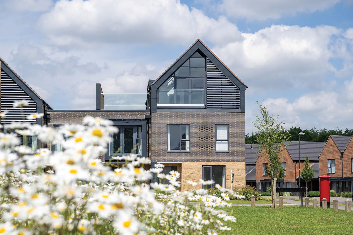 A modern residential neighbourhood featuring a house with a brick and glass exterior, large windows, and a balcony. White flowers are in the foreground, along with a grassy area. Similar houses appear in the background under a partly cloudy sky.