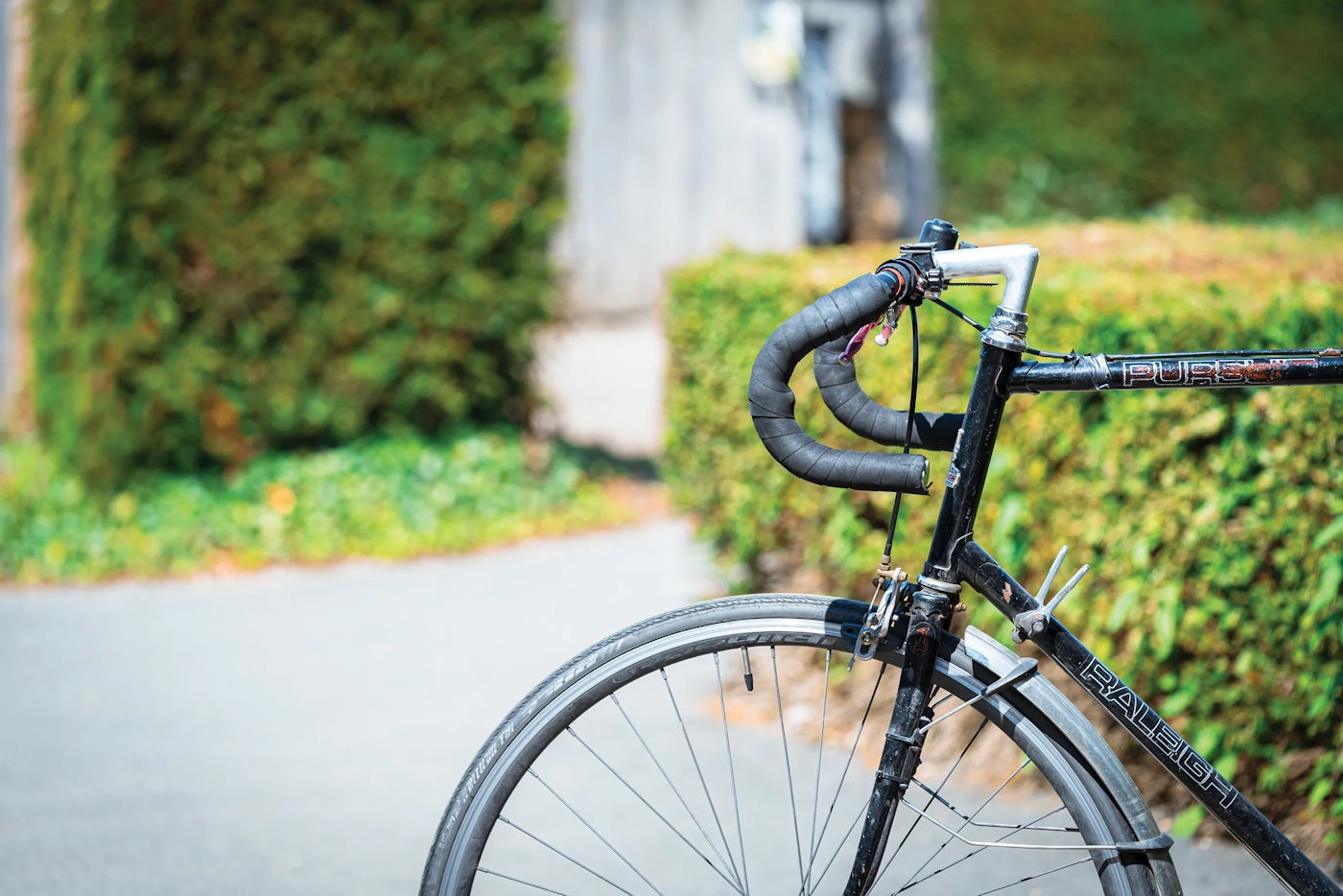 A black Raleigh bicycle is shown in close-up, focusing on the handlebars, front wheel, and part of the frame. The handlebars are wrapped, with brake cables running along them. The background features a paved path lined with green hedges, suggesting an outdoor setting.
