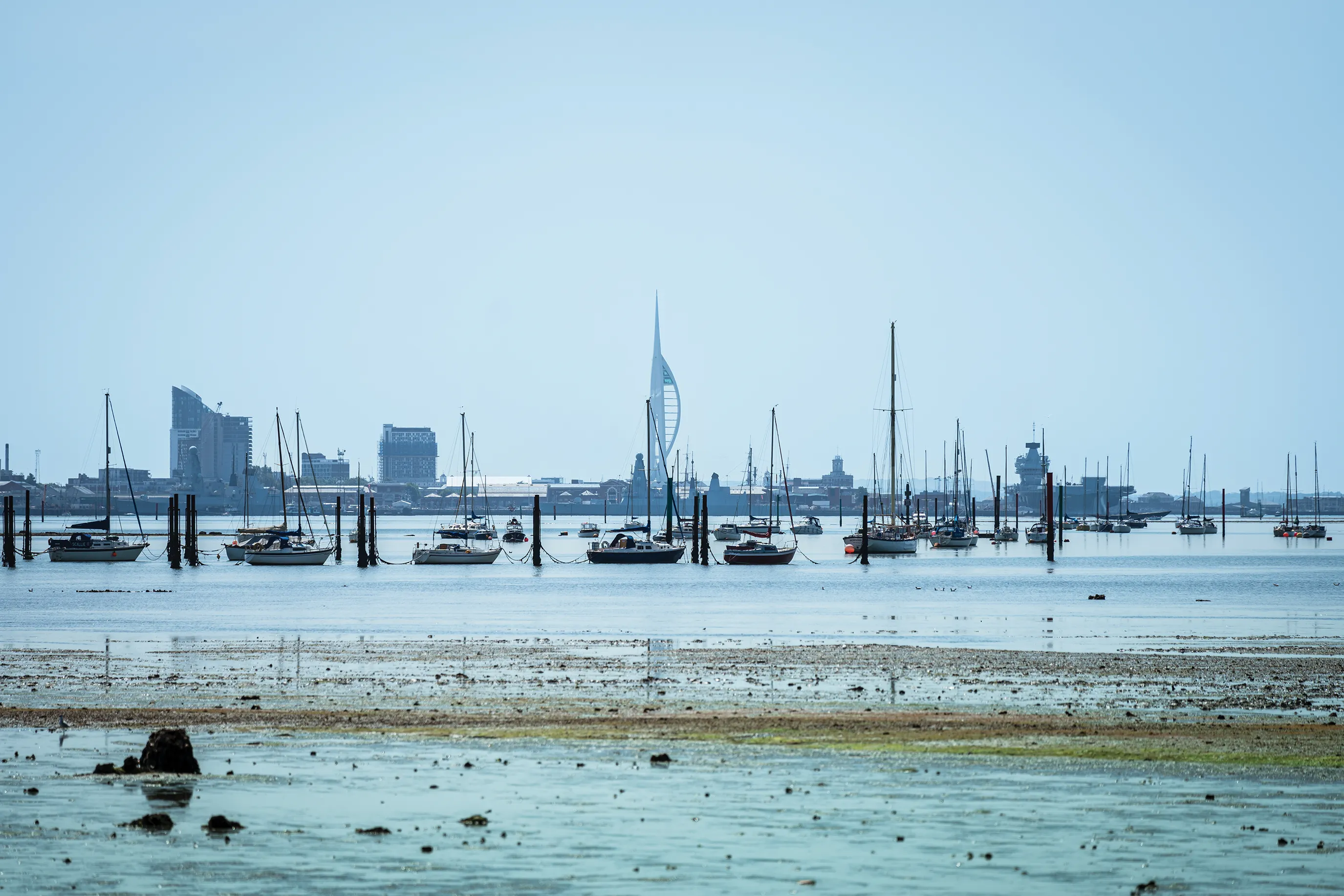 A coastal scene featuring several sailboats anchored in calm waters. In the background, a cityscape of Southsea with modern buildings is visible, including a distinctive tall structure resembling a sail. The foreground includes shallow water with sand and rocks.
