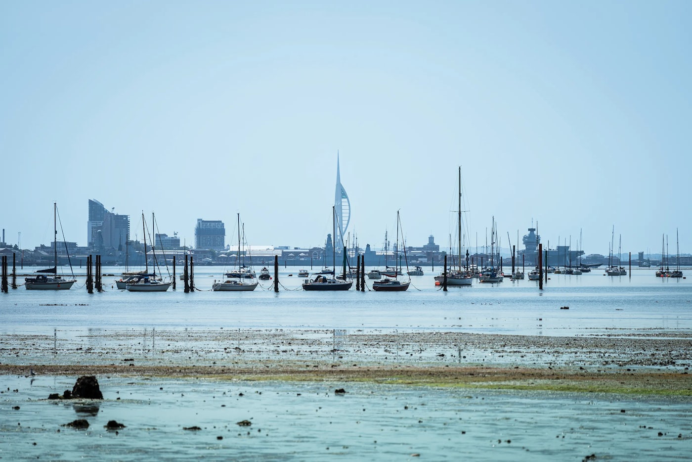 A coastal scene featuring several sailboats anchored in calm waters. In the background, a cityscape of Southsea with modern buildings is visible, including a distinctive tall structure resembling a sail. The foreground includes shallow water with sand and rocks.