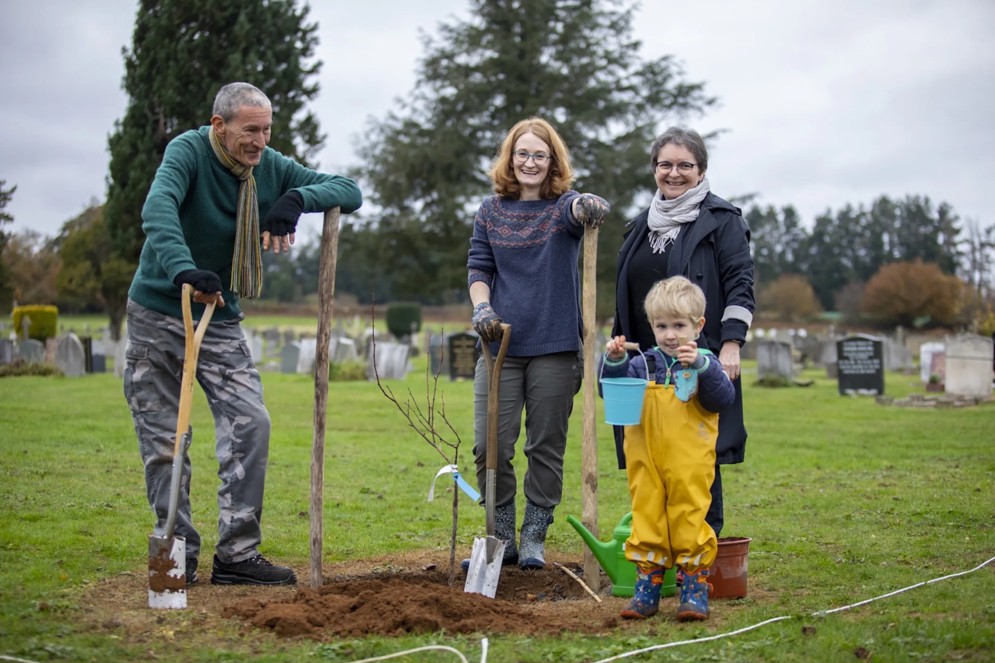Four people are standing in a grassy cemetery, gathered around a newly planted tree. They are holding gardening tools, including shovels and a watering can, indicating they are participating in a tree planting activity. Gravestones are visible in the background, reinforcing the setting as a cemetery.