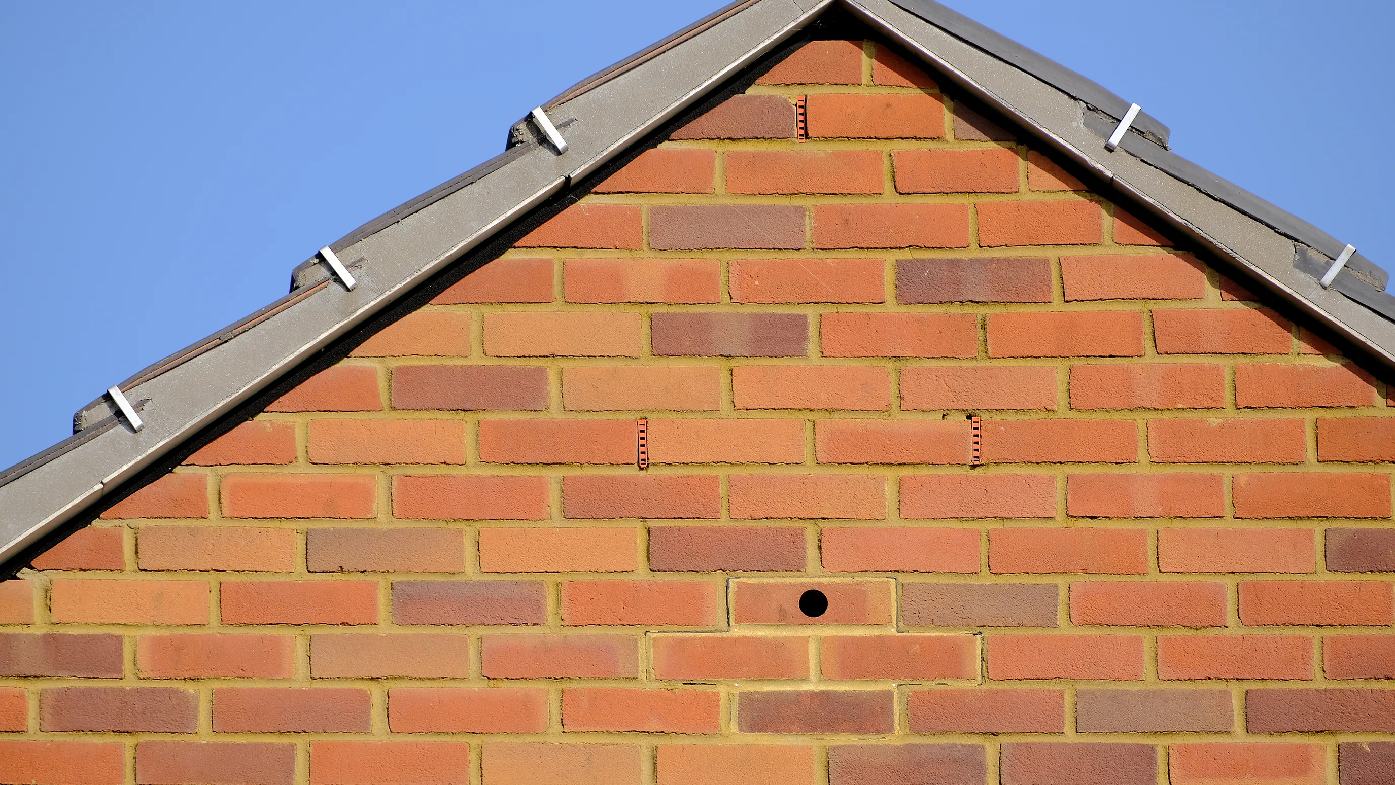 A gable end of a brick house with a sloped roof. The bricks are arranged in a standard pattern. Near the roofline, there is a small circular hole in the brick wall, possibly an entry point for birds or other small animals.
