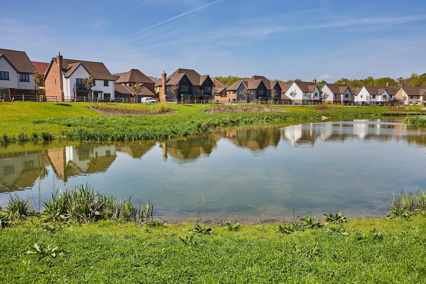 A row of modern houses lines the edge of a calm pond, their facades reflecting in the water. The homes showcase varied architectural styles and colors, including white, brick, and dark-colored exteriors. The foreground features green grass and plants near the water’s edge, enhancing the natural setting.
