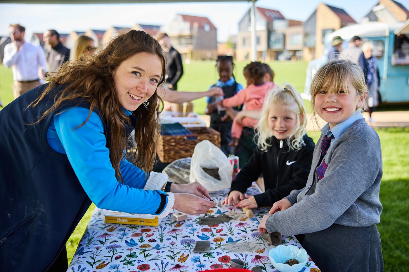 A group of people, including children and adults, are gathered around a table outdoors, engaging in an activity. The table is covered with a floral-patterned tablecloth and holds various items such as leaves, rocks, and a box labeled "Bees." An adult is assisting the children with the activity. The background features other people and buildings, suggesting a community event or gathering.