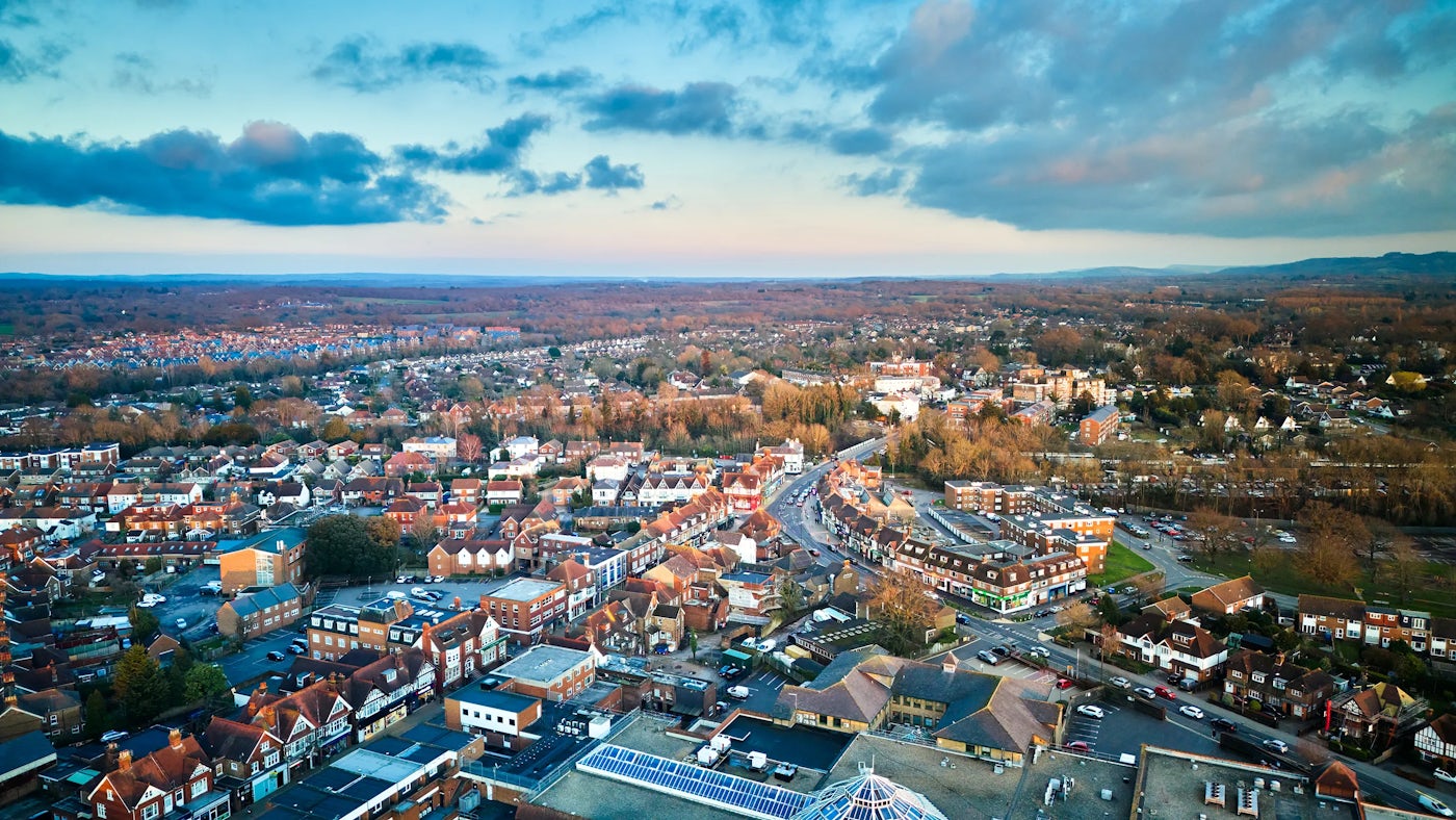 Aerial view of a town with residential and commercial buildings, surrounded by expansive greenery. The sky has a mix of blue and orange hues, suggesting sunrise or sunset.