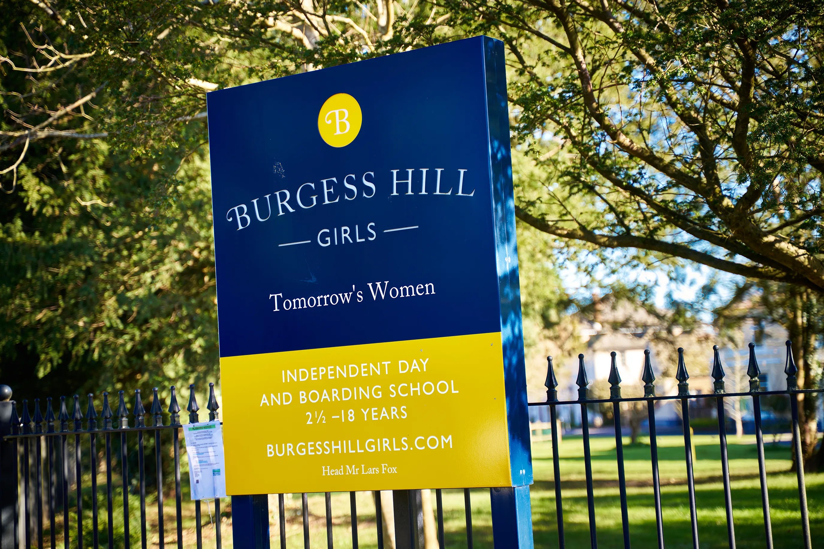 A sign for Burgess Hill Girls, an independent day and boarding school for girls aged 2½ to 18 years. The sign displays the school's name, motto "Tomorrow's Women," and website address (burgesshillgirls.com), with Mr. Lars Fox listed as the head of the school. Positioned in front of a fence, the sign is surrounded by trees and greenery.