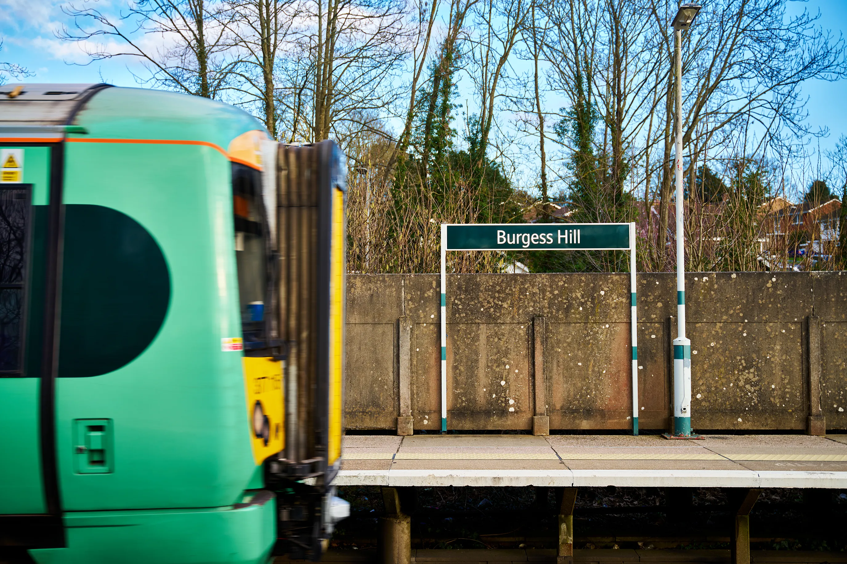 A train station platform with a green train passing by. The platform sign reads "Burgess Hill." Trees and buildings are visible in the background.