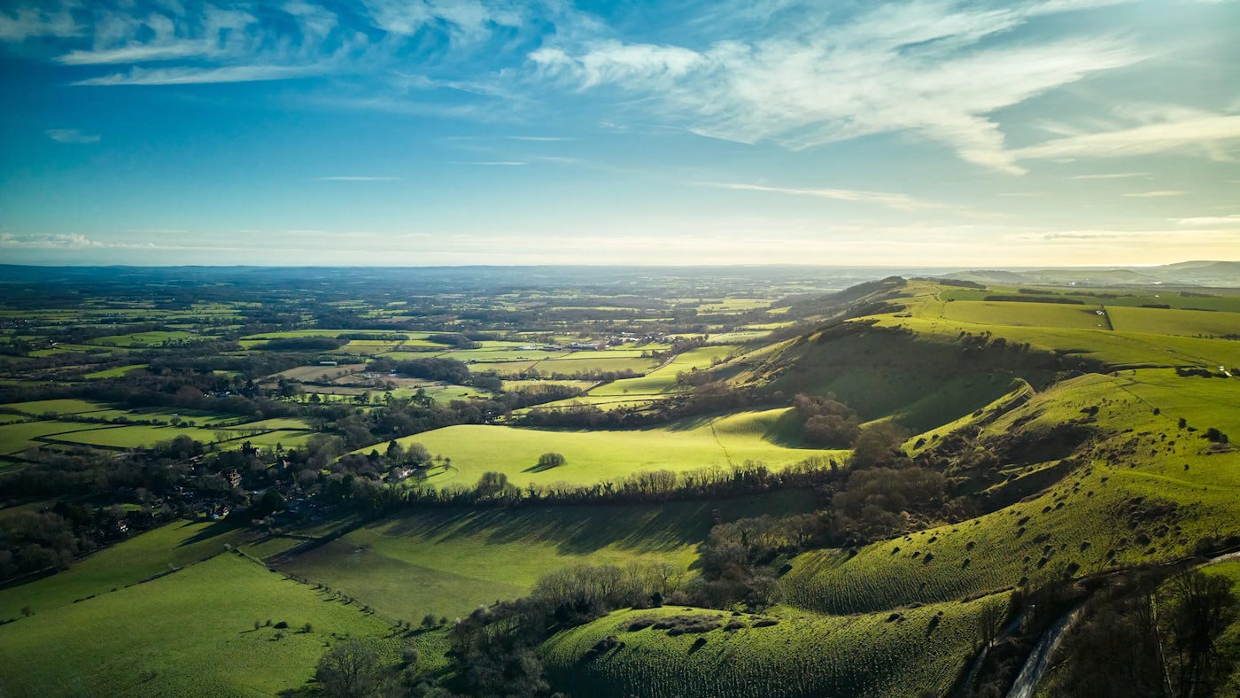 Aerial view of a landscape featuring rolling green hills in the foreground, interspersed with patches of trees and shrubs. Beyond the hills, the terrain transitions into expansive fields with scattered trees. The sky is mostly clear with a few clouds, and the lighting suggests either early morning or late afternoon.