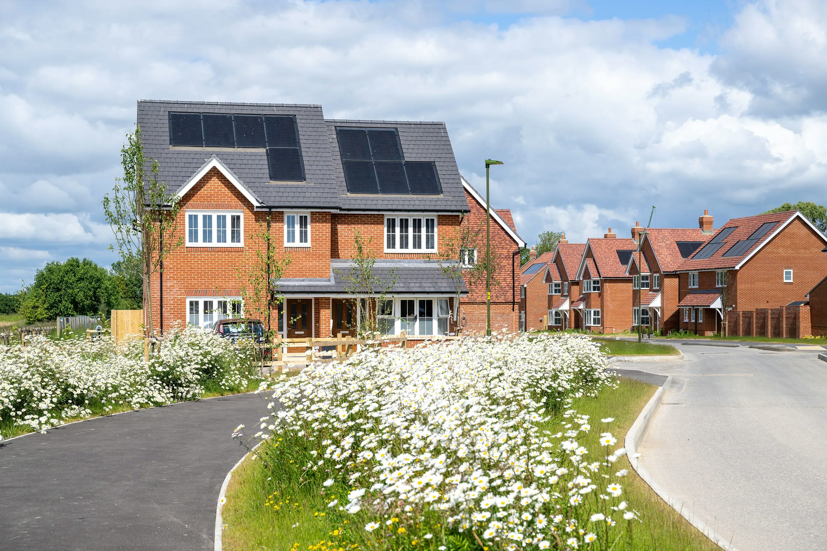 A newly built two-story brick affordable home with solar panels on the roof, surrounded by a landscaped garden with blooming white flowers. A paved walkway leads to the entrance, and a curved road runs alongside the property. In the background, additional houses of similar modern design indicate a newly developed neighbourhood focused on sustainability.