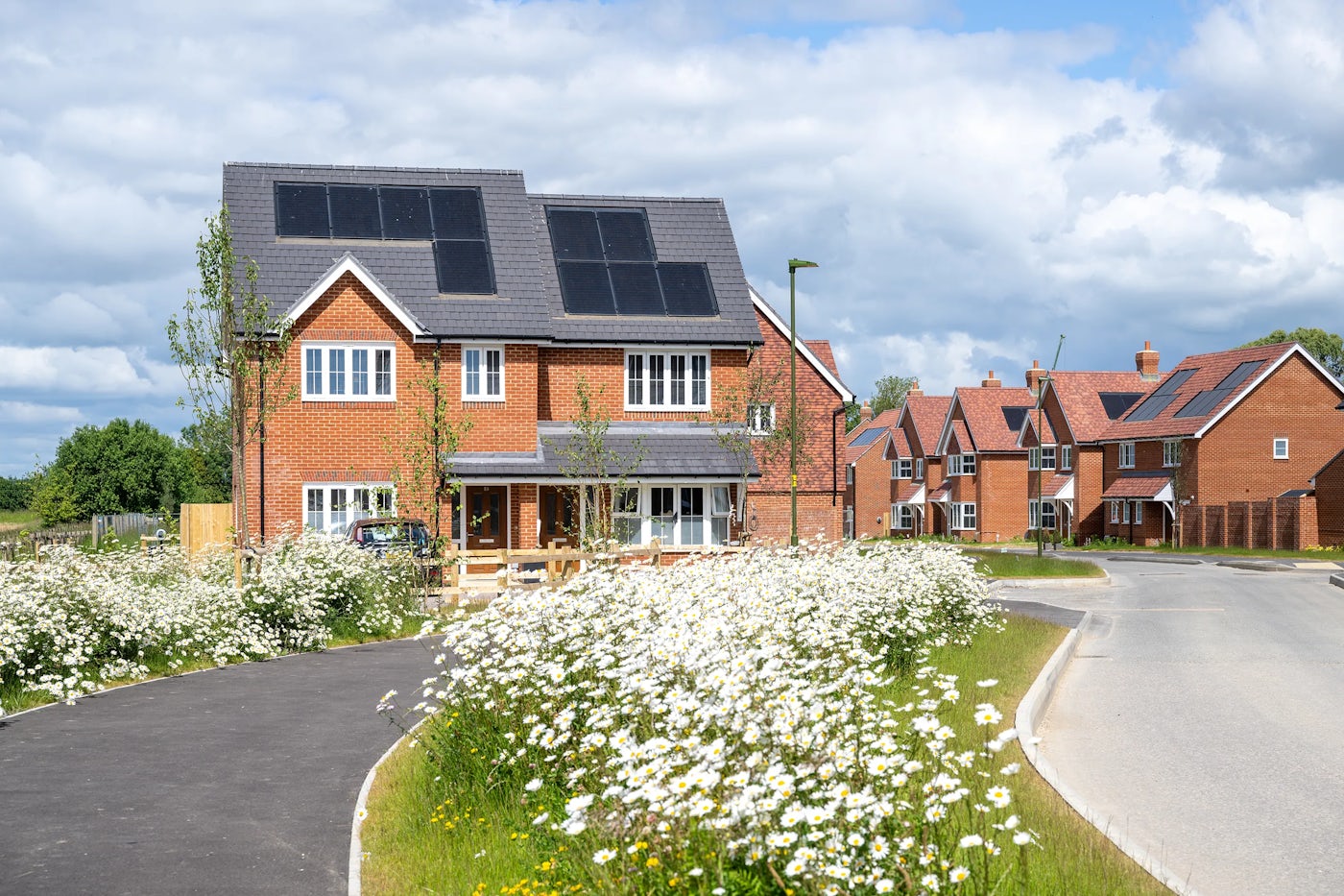 A newly built two-story brick affordable home with solar panels on the roof, surrounded by a landscaped garden with blooming white flowers. A paved walkway leads to the entrance, and a curved road runs alongside the property. In the background, additional houses of similar modern design indicate a newly developed neighbourhood focused on sustainability.