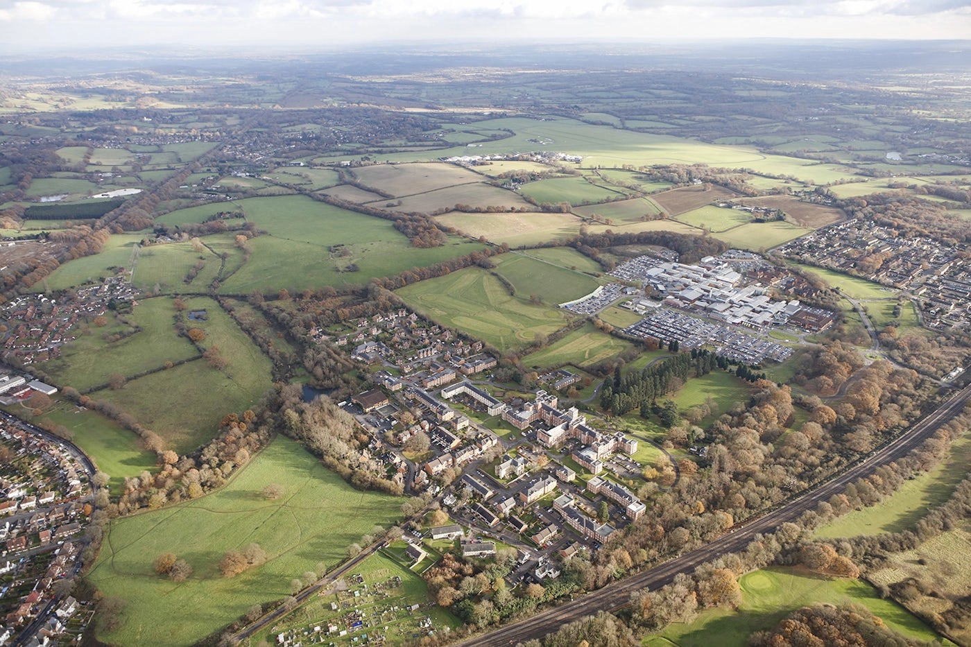 Aerial view of Redhill Hospital and East Surrey Hospital with a mix of residential and commercial buildings, open green fields, and clusters of trees. Roads and pathways connect different areas, and small bodies of water are visible throughout the terrain.