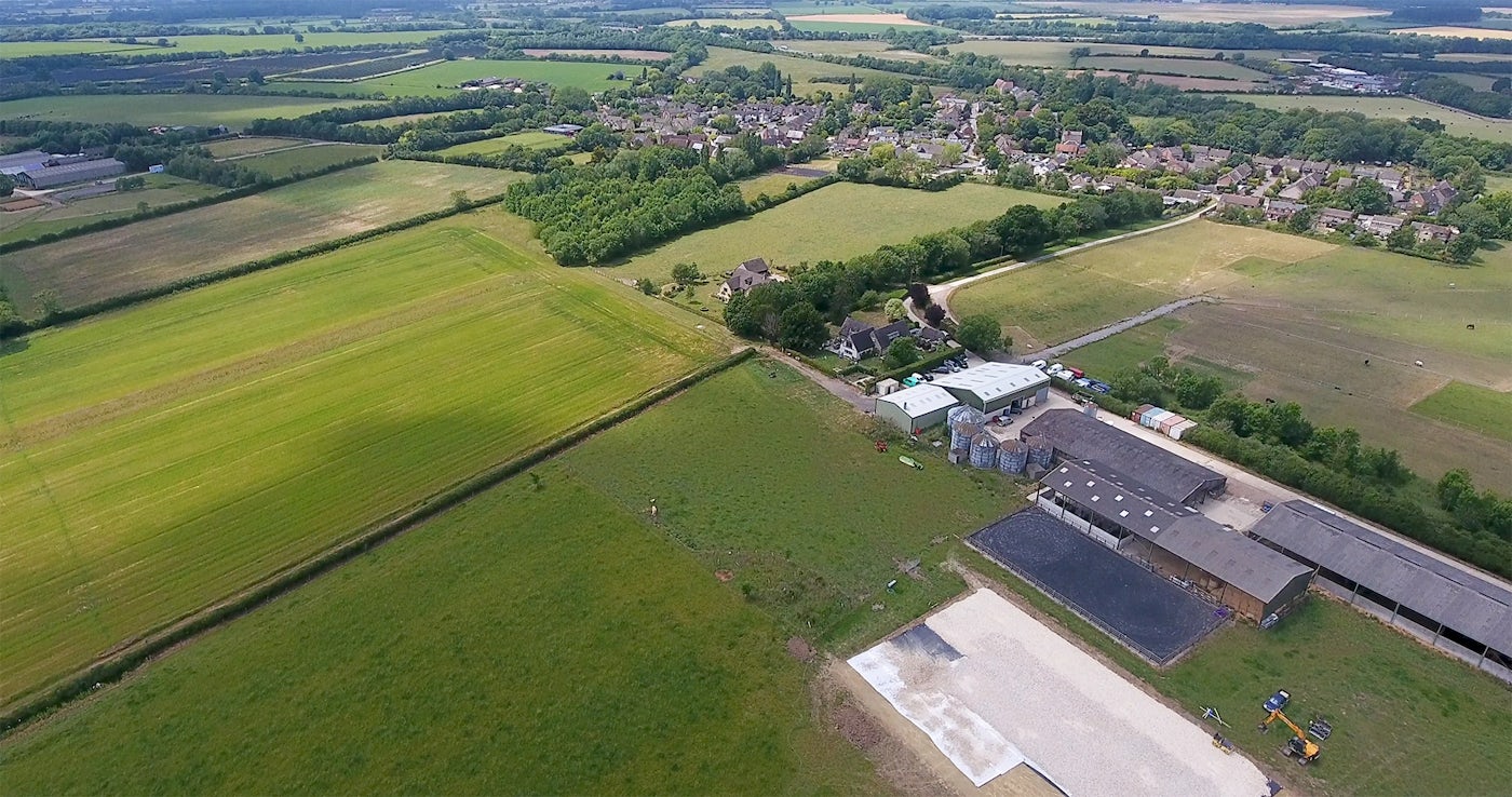 Aerial view of a rural landscape at Wendlebury in South Bicester featuring agricultural fields, farm buildings, and a small village. The foreground showcases fields in varying shades of green and brown, indicating different crops or growth stages. Several farm structures, including barns and silos, are visible, along with scattered machinery. In the background, a small village with houses surrounded by trees is nestled among more fields extending into the horizon.