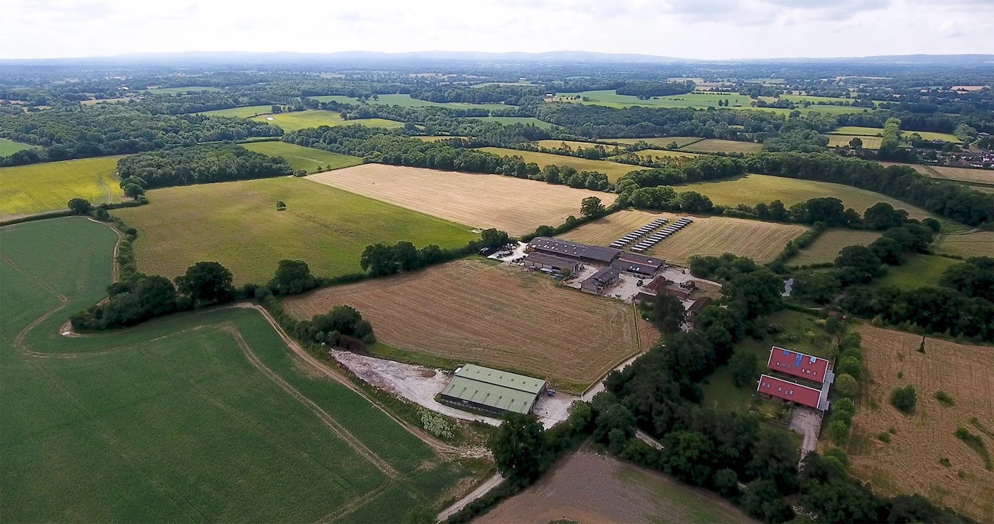 Aerial view of a rural area of West Grinstead with expansive fields in varying shades of green and brown, a small village, and several farm buildings. The village is surrounded by trees, with houses interspersed among them. The farm buildings include silos, barns, and other agricultural structures, with vehicles and equipment visible nearby.