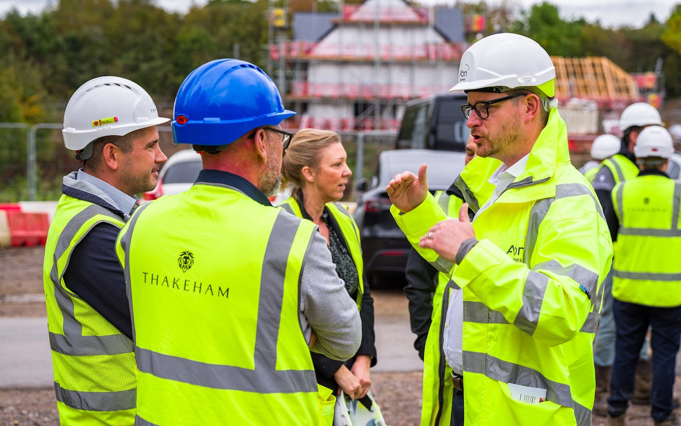 Group of people at a construction site wearing high-visibility jackets and hard hats. One person in the foreground has "THAKEHAM" written on the back of their jacket. The group appears engaged in a discussion, with one person wearing a blue hard hat while the others wear white hard hats. The background features construction materials and scaffolding, indicating an active building project.
