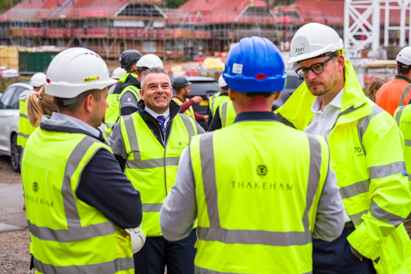 Group of people at a construction site wearing high-visibility jackets and hard hats, engaged in a discussion. One person in the foreground has "THAKEHAM" printed on the back of their jacket. The group includes individuals with white and blue hard hats. Scaffolding and construction materials are visible in the background, indicating an active building site.