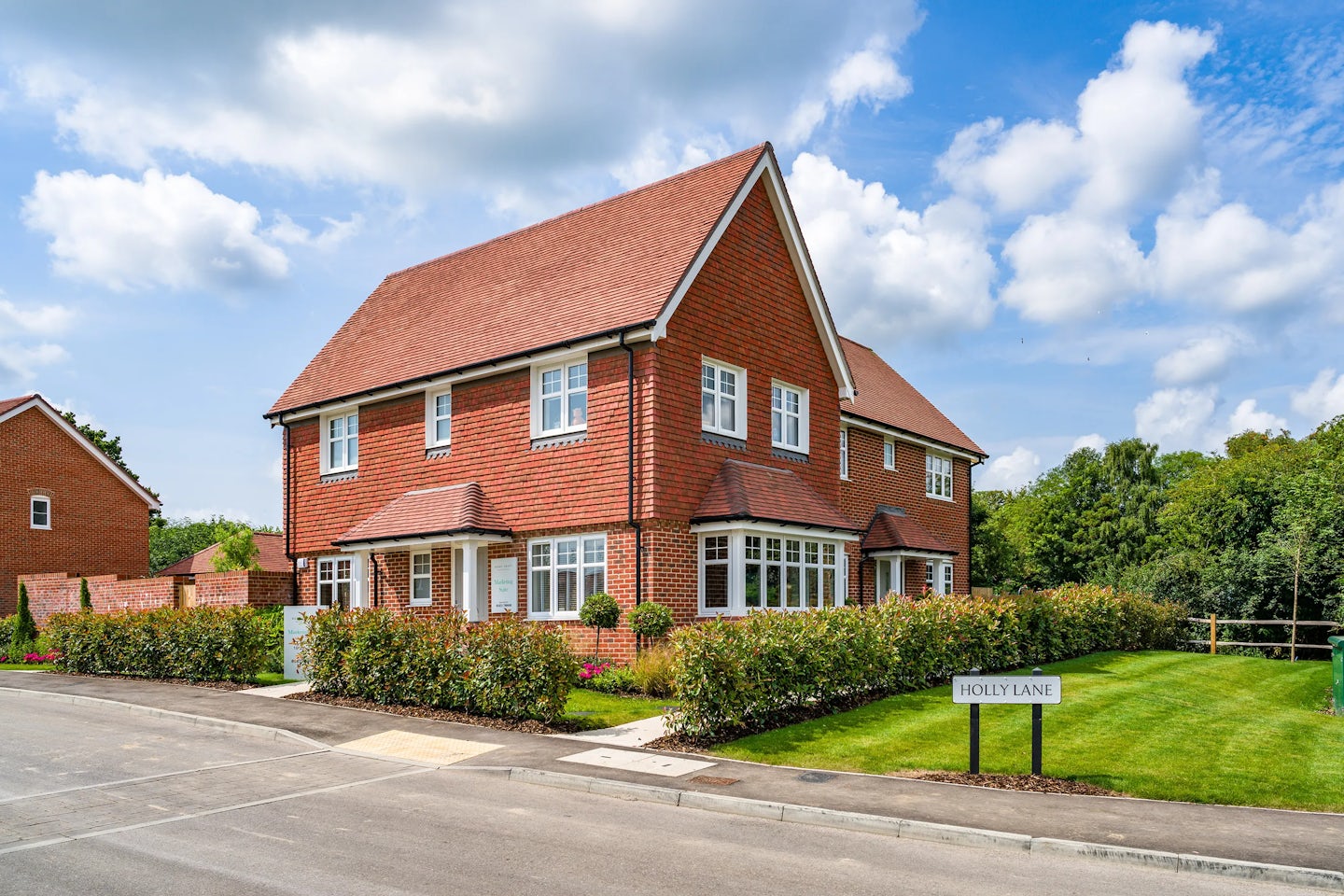 A modern, two-story brick house with a red-tiled roof on a corner lot. The house features multiple windows, a well-maintained garden with shrubs, and a driveway. In the foreground, a street sign reads "HOLLY LANE." The sky is partly cloudy with patches of blue.
