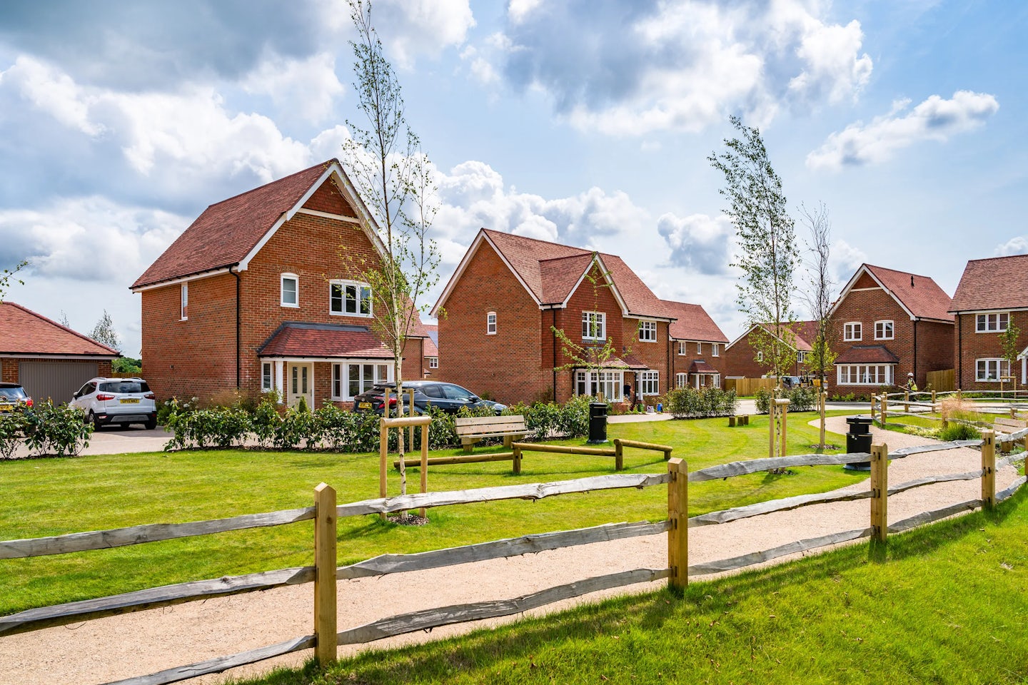 A suburban neighborhood with modern brick houses featuring red-tiled roofs. The houses are surrounded by well-maintained lawns, trees, and shrubs. A paved pathway with wooden fencing runs through the green space in front of the houses, and several cars are parked in the driveways. The sky is partly cloudy.