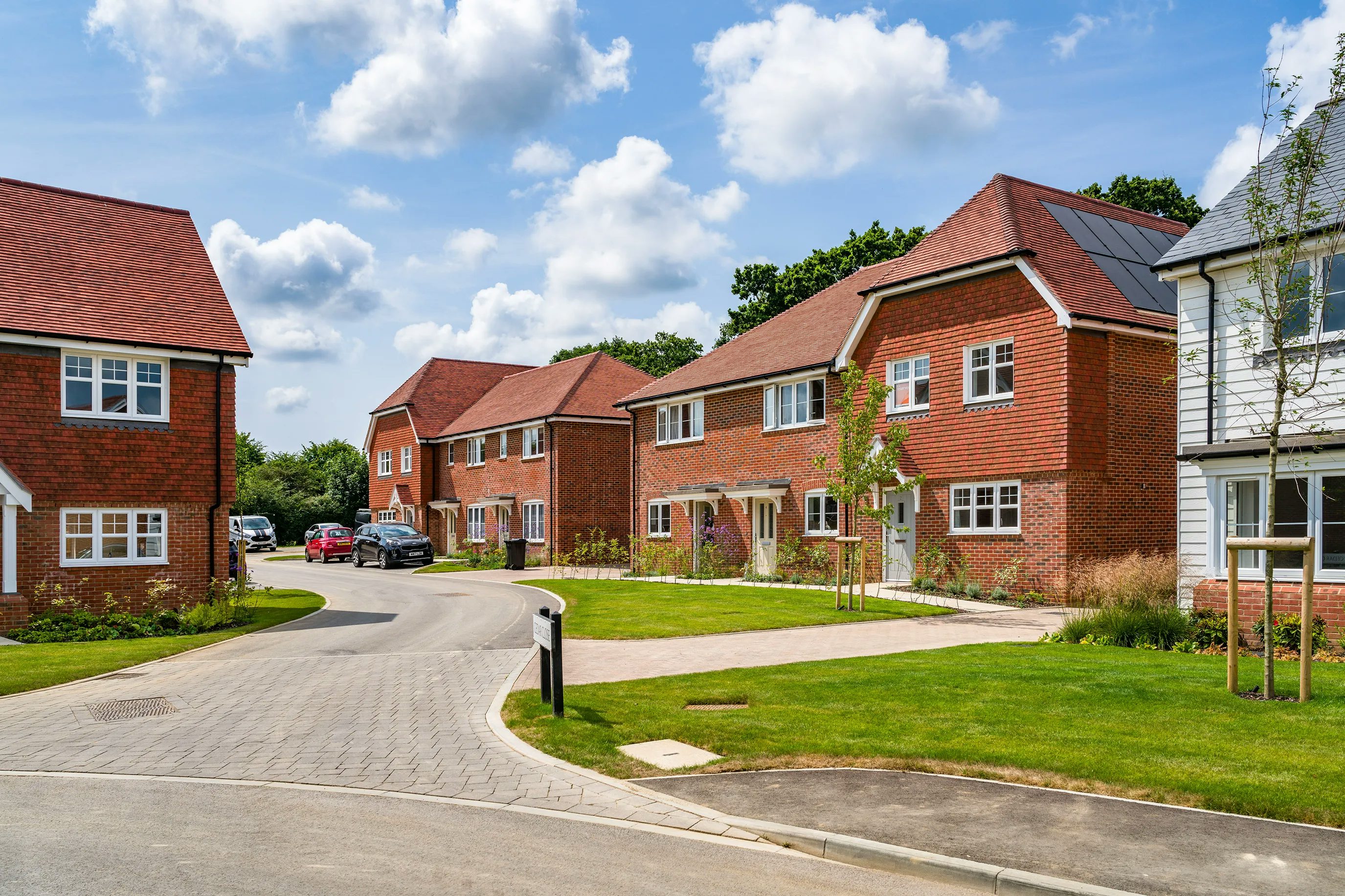 A suburban neighborhood with modern, detached houses featuring red brick exteriors and pitched roofs, some with solar panels. The curved, paved street is lined with well-maintained lawns and small trees. A few cars are parked along the street. The sky is blue with scattered clouds, indicating a pleasant day.