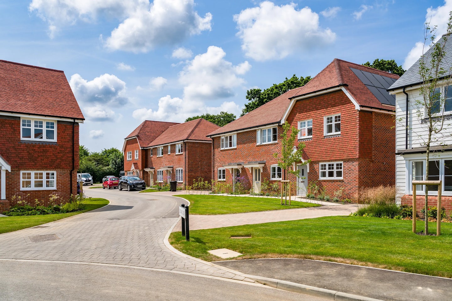 A suburban neighborhood with modern, detached houses featuring red brick exteriors and pitched roofs, some with solar panels. The curved, paved street is lined with well-maintained lawns and small trees. A few cars are parked along the street. The sky is blue with scattered clouds, indicating a pleasant day.