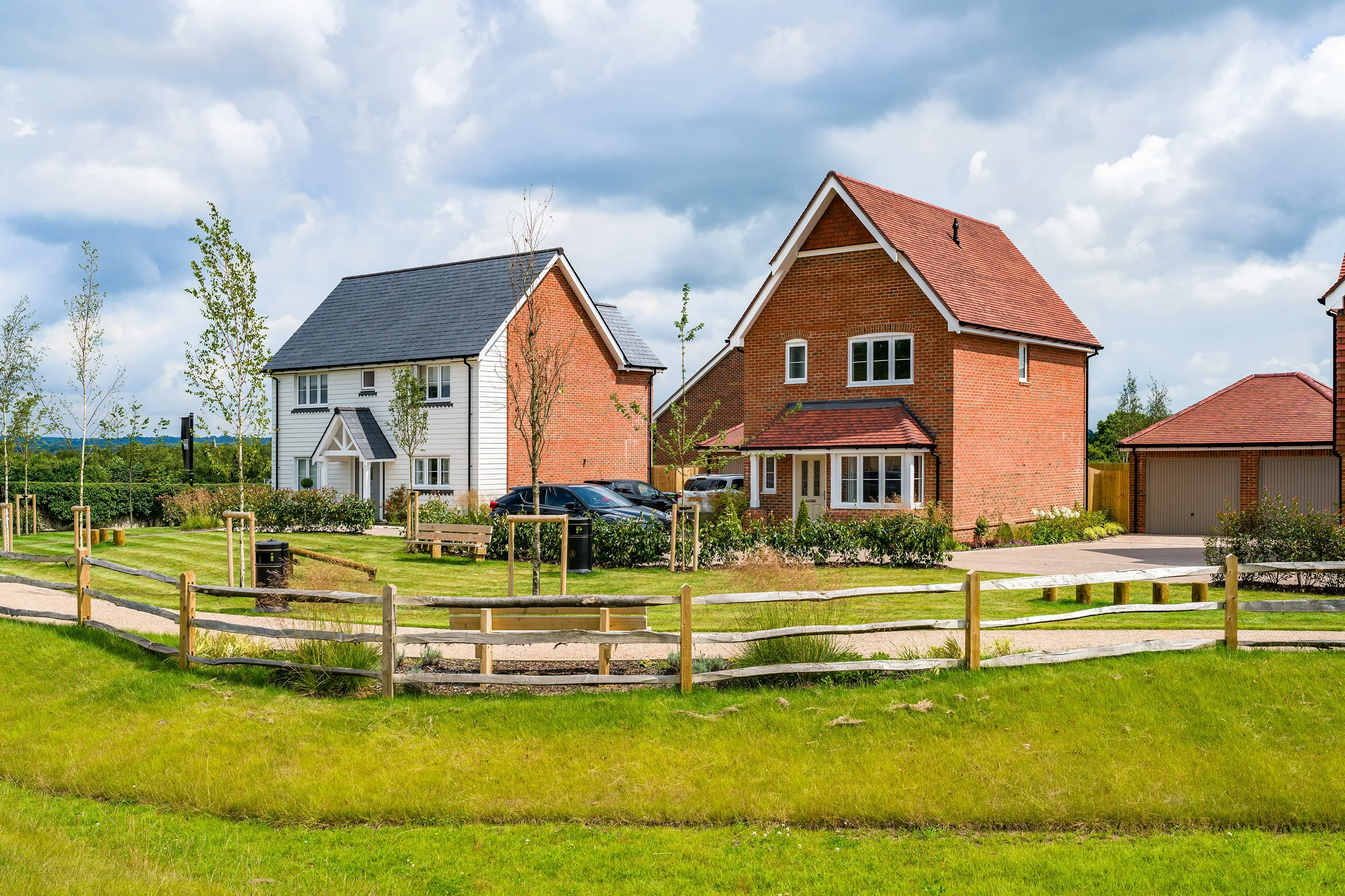 Two modern suburban houses, one white with a dark grey roof and the other red brick with a red roof, stand side by side. Both have well-maintained gardens and driveways. A wooden fence in the foreground separates a grassy area from the houses. The sky is partly cloudy.