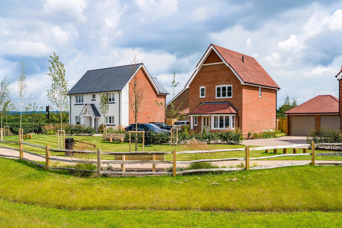 Two modern suburban houses, one white with a dark grey roof and the other red brick with a red roof, stand side by side. Both have well-maintained gardens and driveways. A wooden fence in the foreground separates a grassy area from the houses. The sky is partly cloudy.