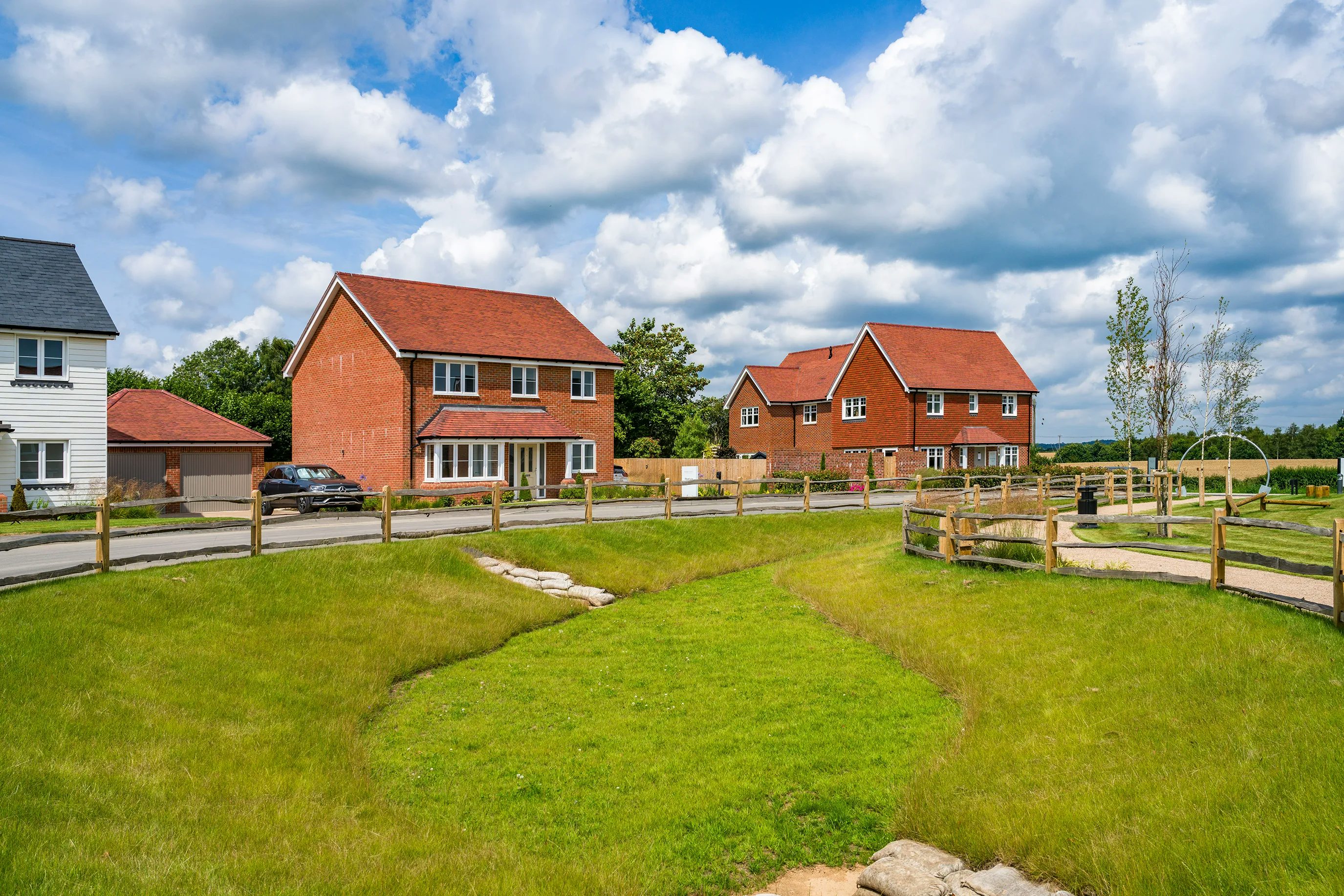 Three suburban houses with red brick exteriors and red-tiled roofs line a curved road. Each house has a well-maintained lawn, and a wooden fence separates the road from a grassy area. The sky is partly cloudy with blue patches visible.