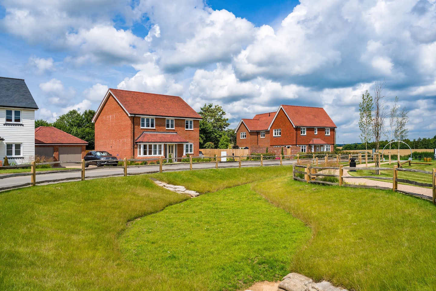 Three suburban houses with red brick exteriors and red-tiled roofs line a curved road. Each house has a well-maintained lawn, and a wooden fence separates the road from a grassy area. The sky is partly cloudy with blue patches visible.