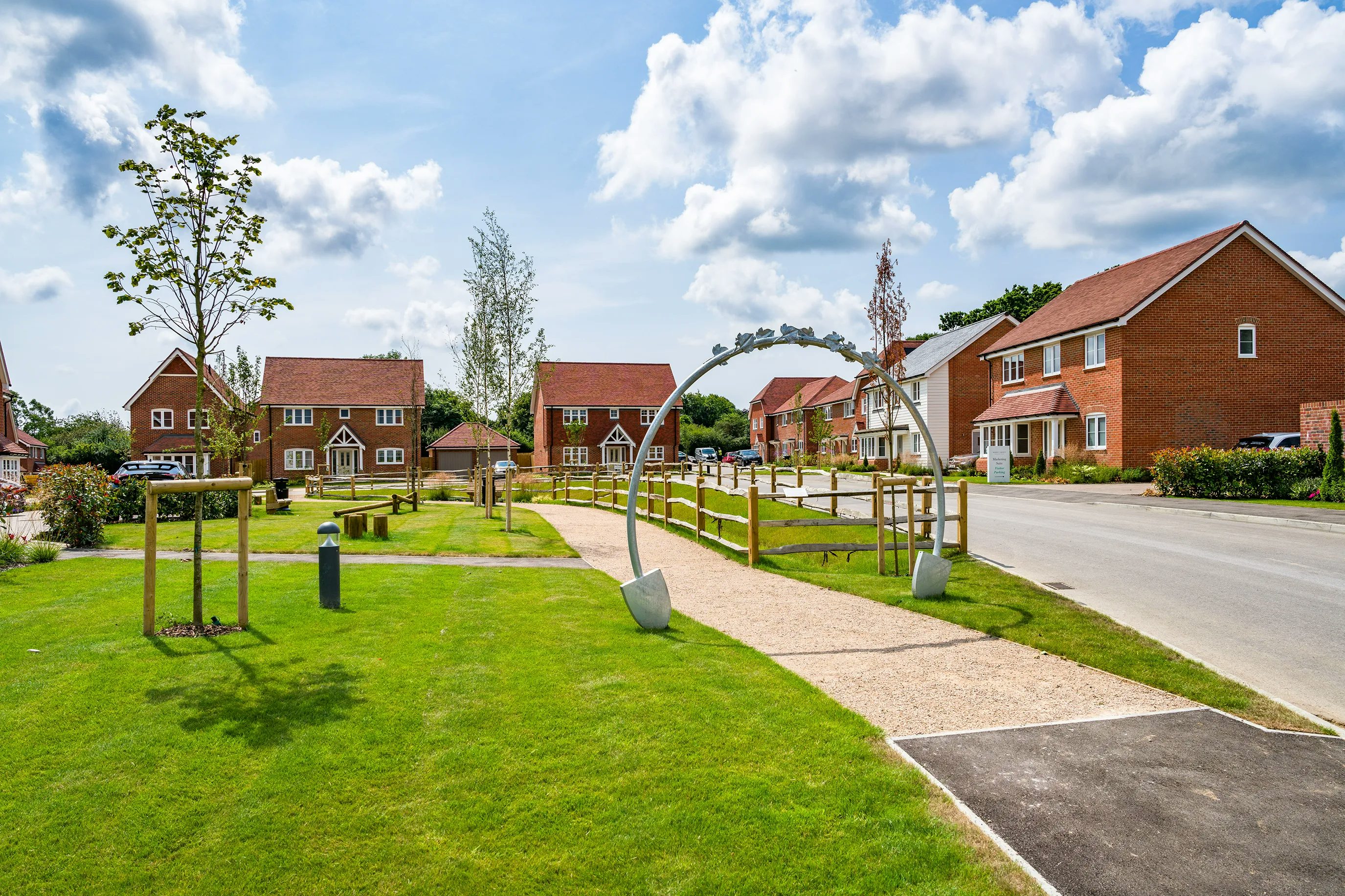 A suburban neighborhood with brick houses featuring red-tiled roofs, green lawns, and young trees. A paved walkway leads through the area, passing under a metal archway. The sky is partly cloudy with patches of blue.