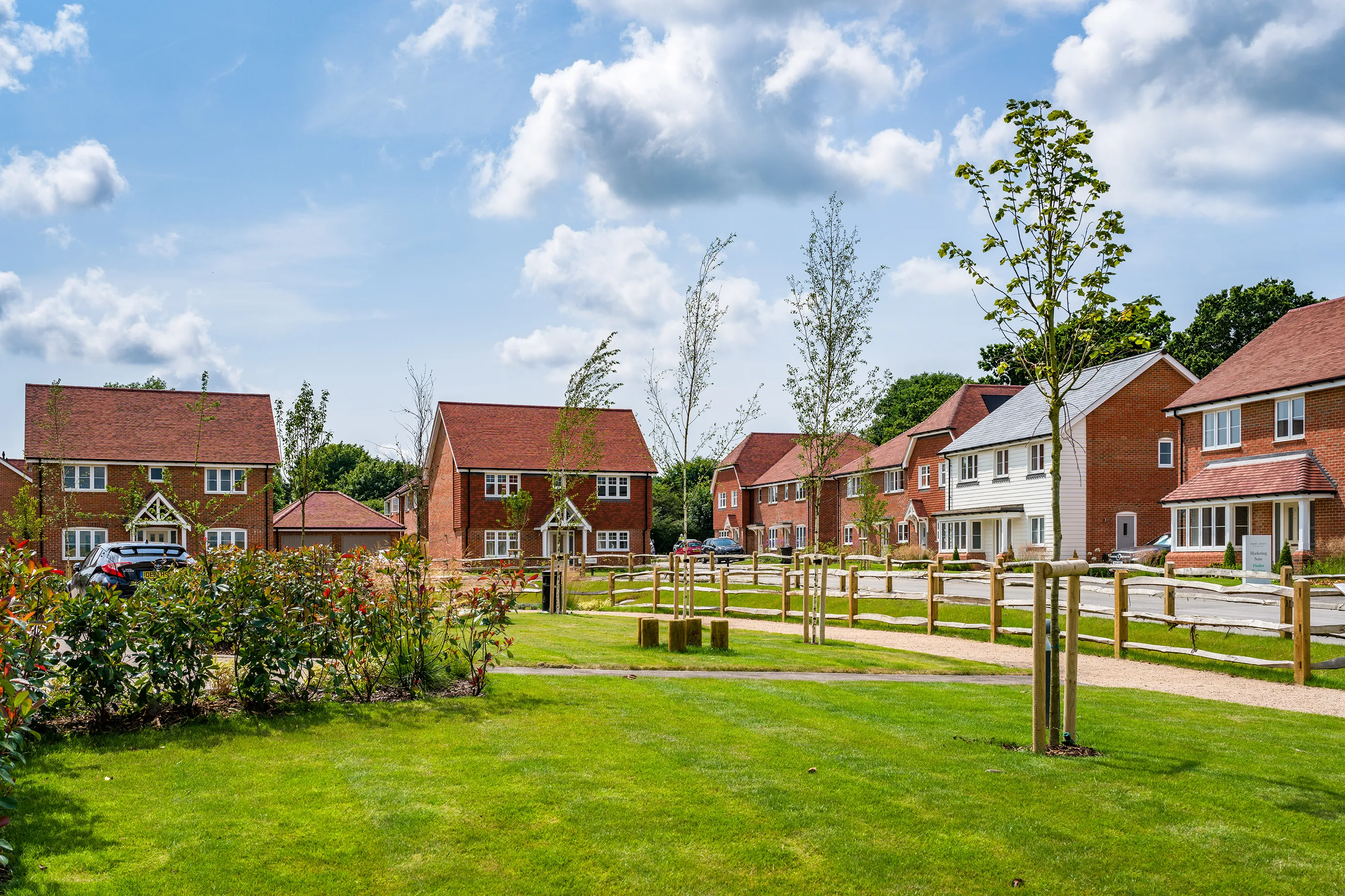 A suburban neighborhood with detached houses featuring red brick exteriors and pitched roofs. The homes are arranged along a street with parked cars visible. In the foreground, a well-maintained grassy area contains young trees and shrubs, enclosed by wooden fencing. The sky is partly cloudy with patches of blue.
