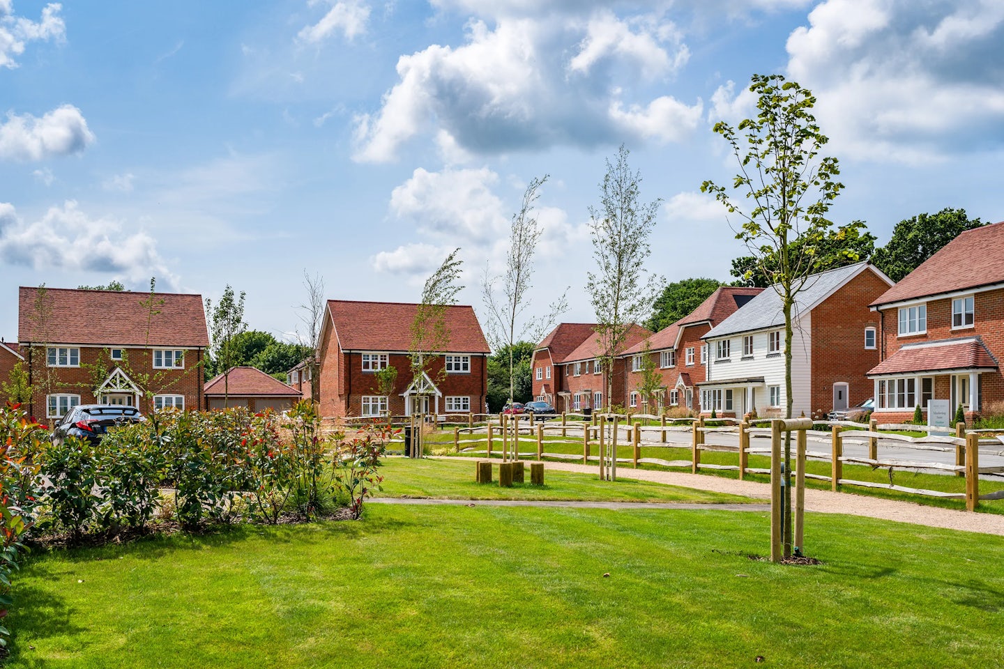 A suburban neighborhood with detached houses featuring red brick exteriors and pitched roofs. The homes are arranged along a street with parked cars visible. In the foreground, a well-maintained grassy area contains young trees and shrubs, enclosed by wooden fencing. The sky is partly cloudy with patches of blue.