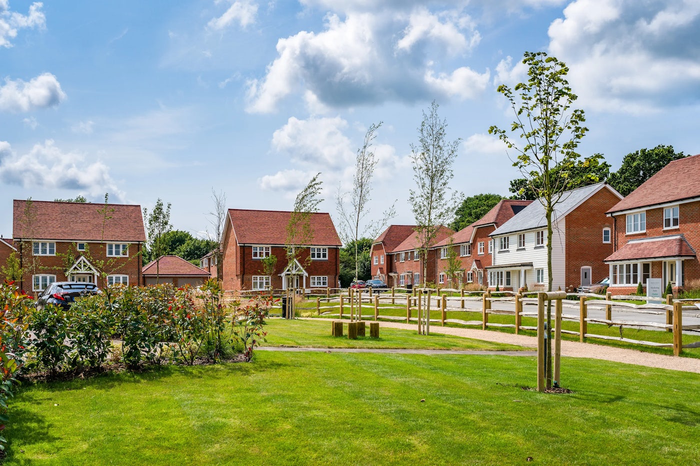 A suburban neighborhood with detached houses featuring red brick exteriors and pitched roofs. The homes are arranged along a street with parked cars visible. In the foreground, a well-maintained grassy area contains young trees and shrubs, enclosed by wooden fencing. The sky is partly cloudy with patches of blue.