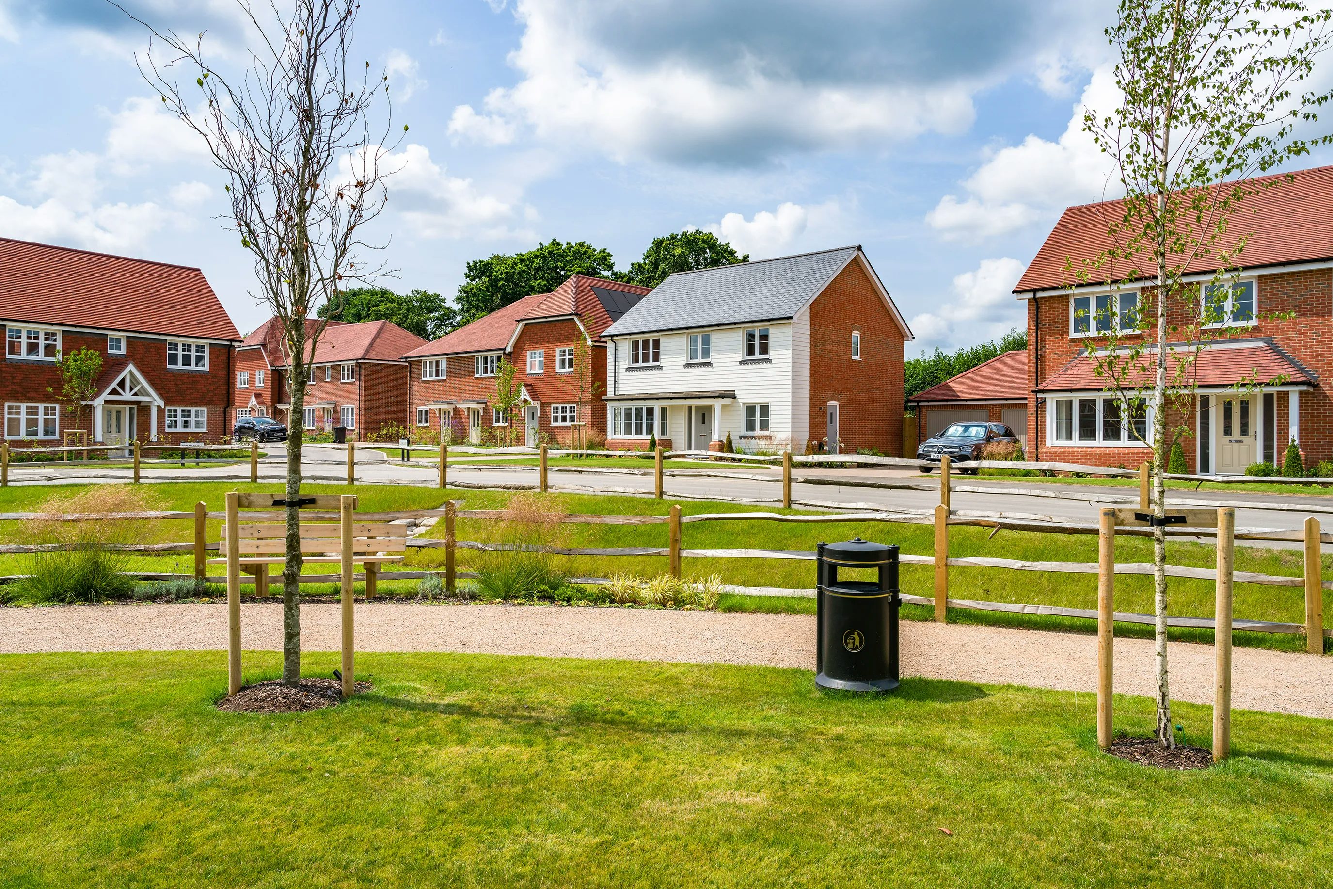 A suburban neighborhood with detached houses featuring pitched roofs and brick facades. A paved road runs through the scene, lined with neatly trimmed hedges and small front gardens. The houses have driveways, some with parked cars. The sky is partly cloudy, casting soft shadows on the buildings and street.