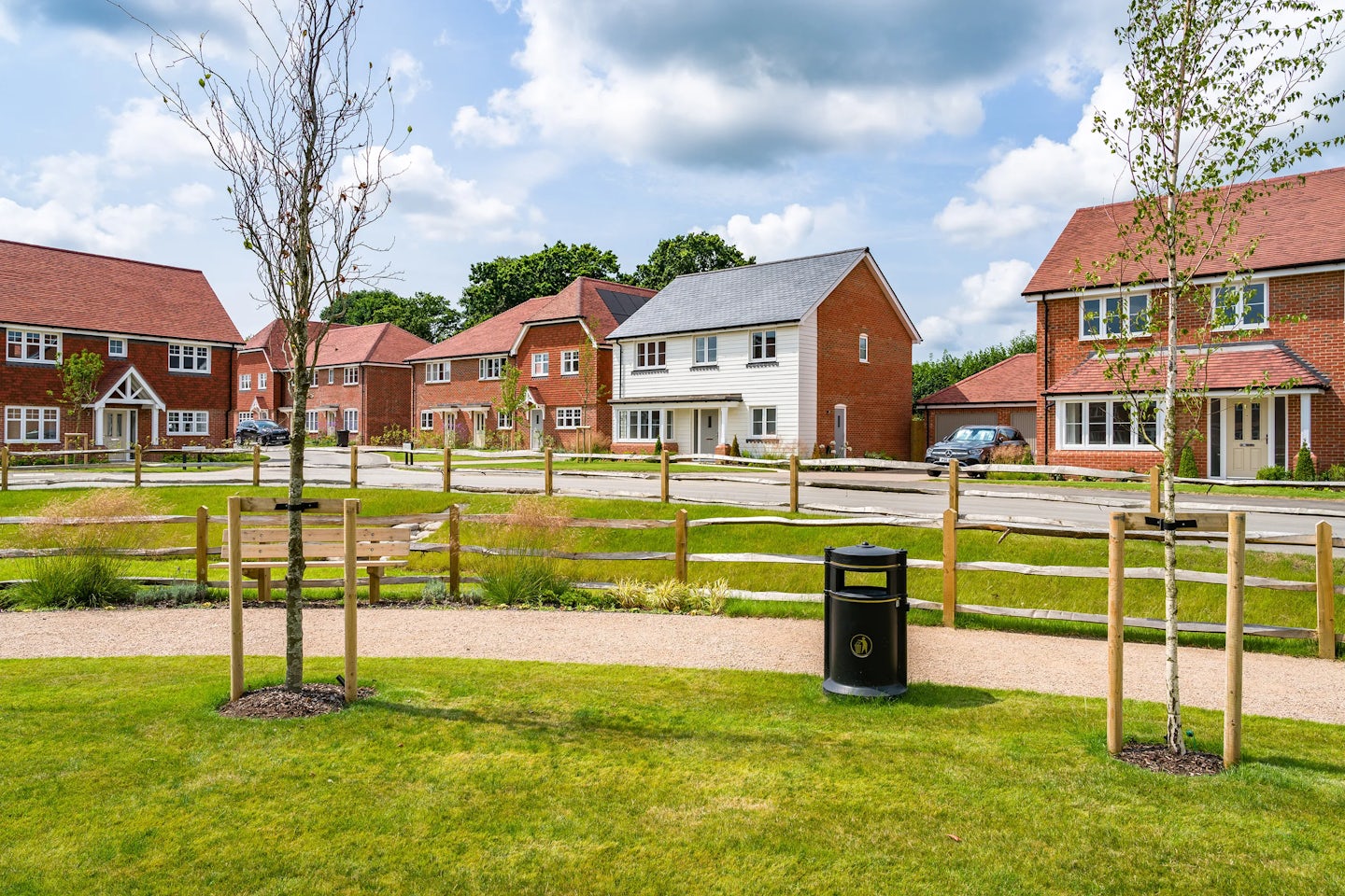 A suburban neighborhood with detached houses featuring pitched roofs and brick facades. A paved road runs through the scene, lined with neatly trimmed hedges and small front gardens. The houses have driveways, some with parked cars. The sky is partly cloudy, casting soft shadows on the buildings and street.