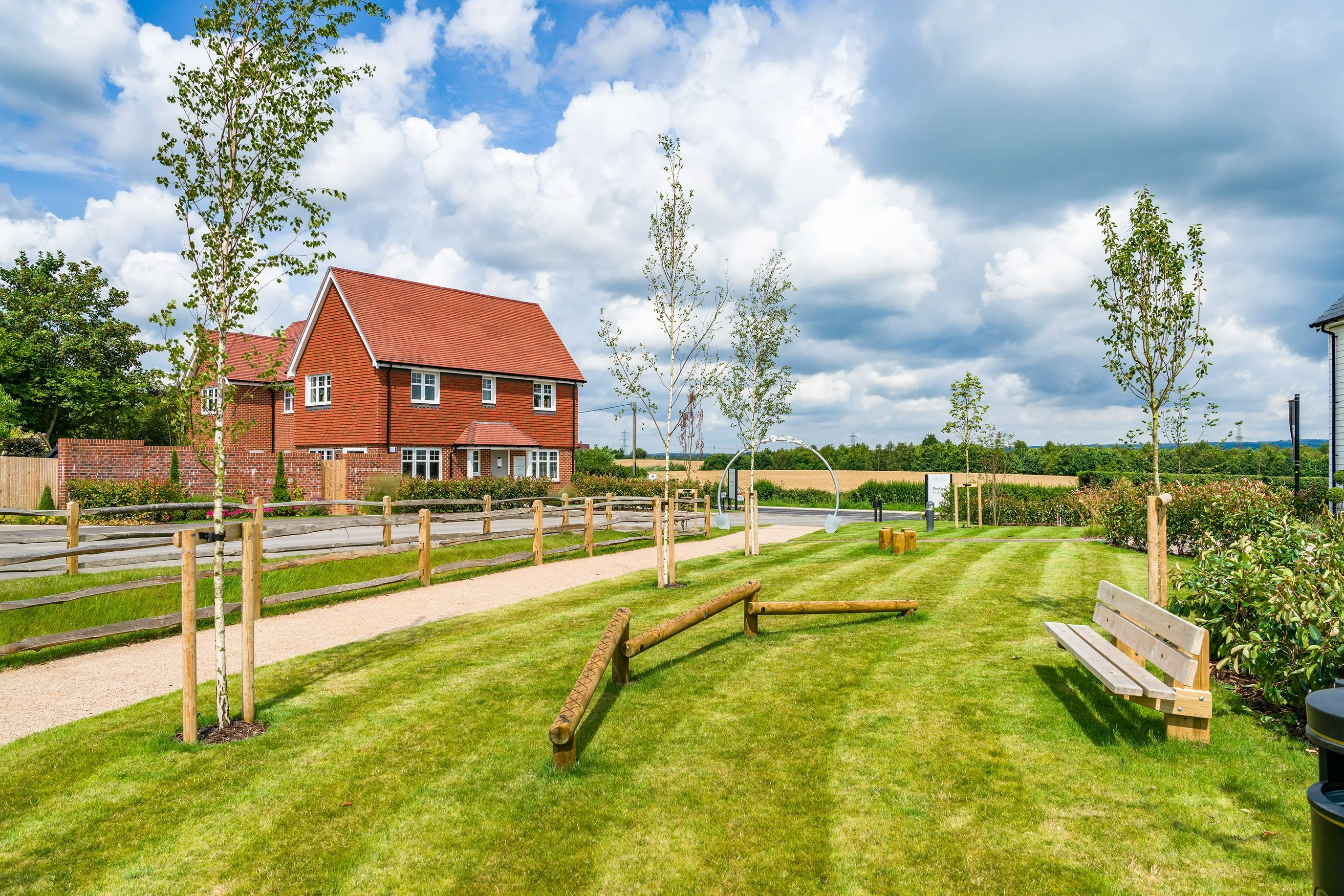 A landscaped outdoor area with a red-brick house in the background. The scene includes a well-maintained lawn, young trees with wooden stakes for support, a gravel pathway, wooden benches, and logs arranged on the grass. The sky is partly cloudy, and the setting is rural.