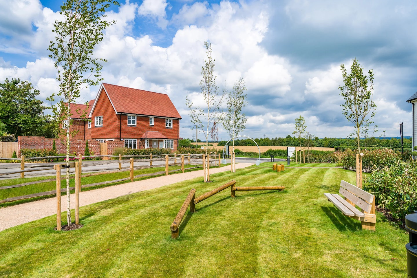 A landscaped outdoor area with a red-brick house in the background. The scene includes a well-maintained lawn, young trees with wooden stakes for support, a gravel pathway, wooden benches, and logs arranged on the grass. The sky is partly cloudy, and the setting is rural.
