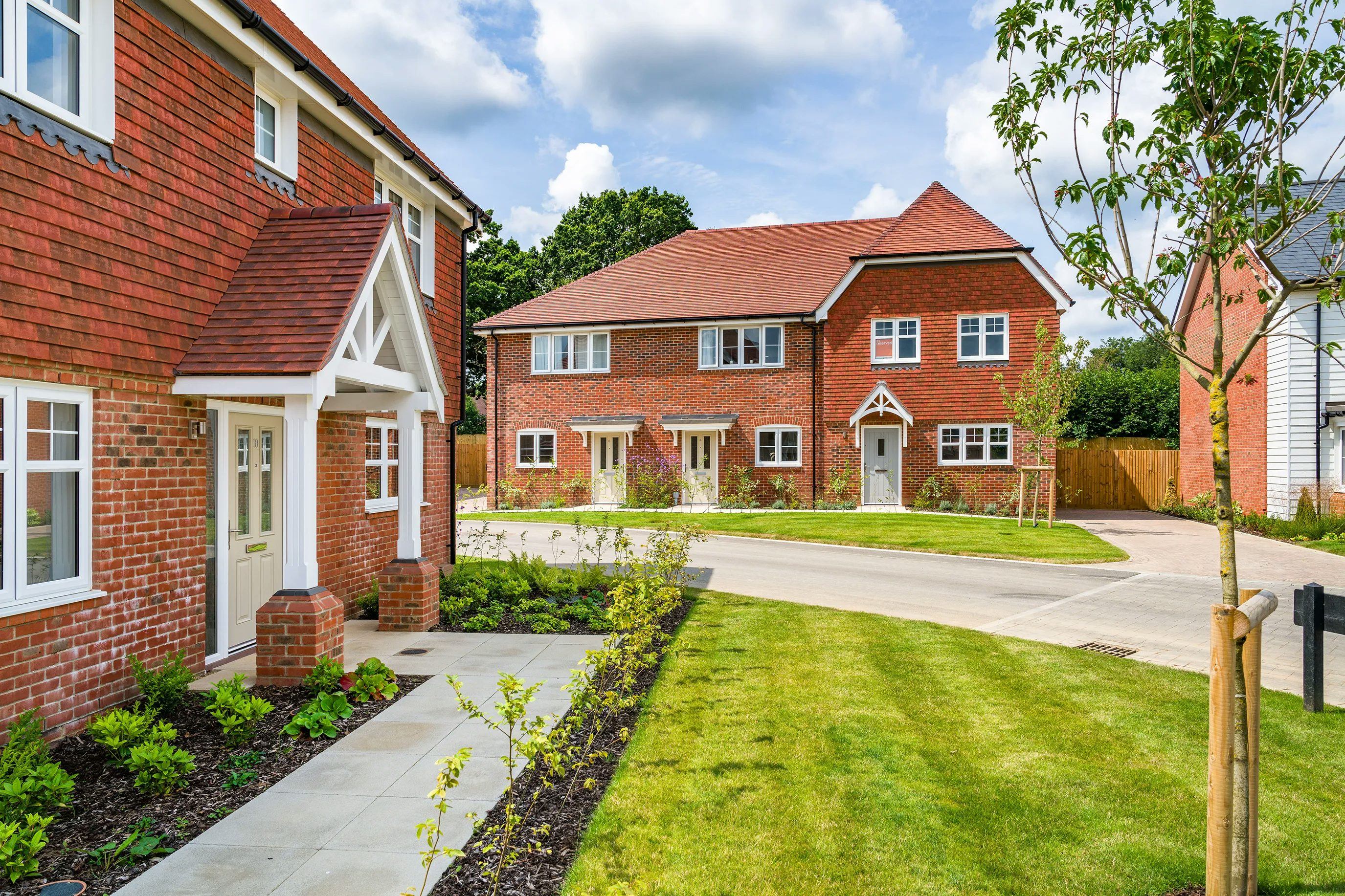 A row of newly built houses constructed with red brick, featuring white-framed windows and doors. The house on the left includes a porch with a triangular roof, while the duplex in the background has two front doors with small gardens in front. The area is landscaped with green lawns, young trees, and shrubs. The sky is partly cloudy, and the overall scene is bright and well-maintained.