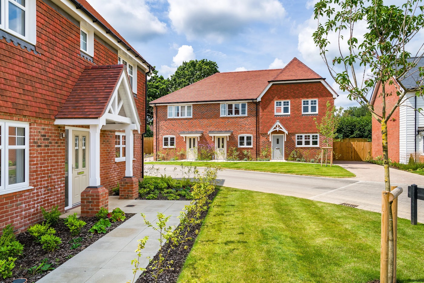 A row of newly built houses constructed with red brick, featuring white-framed windows and doors. The house on the left includes a porch with a triangular roof, while the duplex in the background has two front doors with small gardens in front. The area is landscaped with green lawns, young trees, and shrubs. The sky is partly cloudy, and the overall scene is bright and well-maintained.