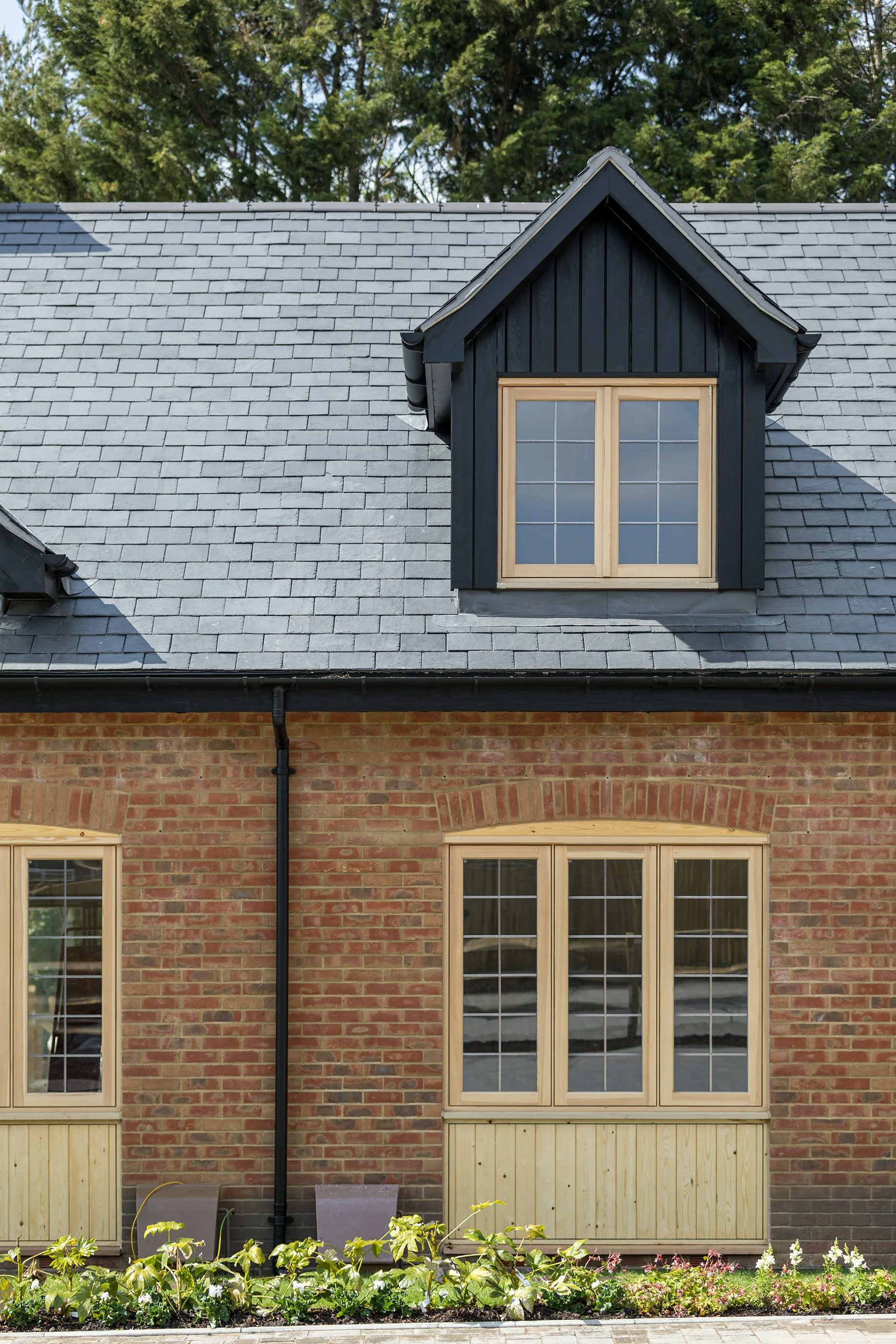 A brick house with a slate roof, featuring a dormer window framed in dark wood with two panes. Below, two ground-floor windows have multiple panes and wooden frames. The house is surrounded by greenery, including trees in the background and a garden with flowers in the foreground.