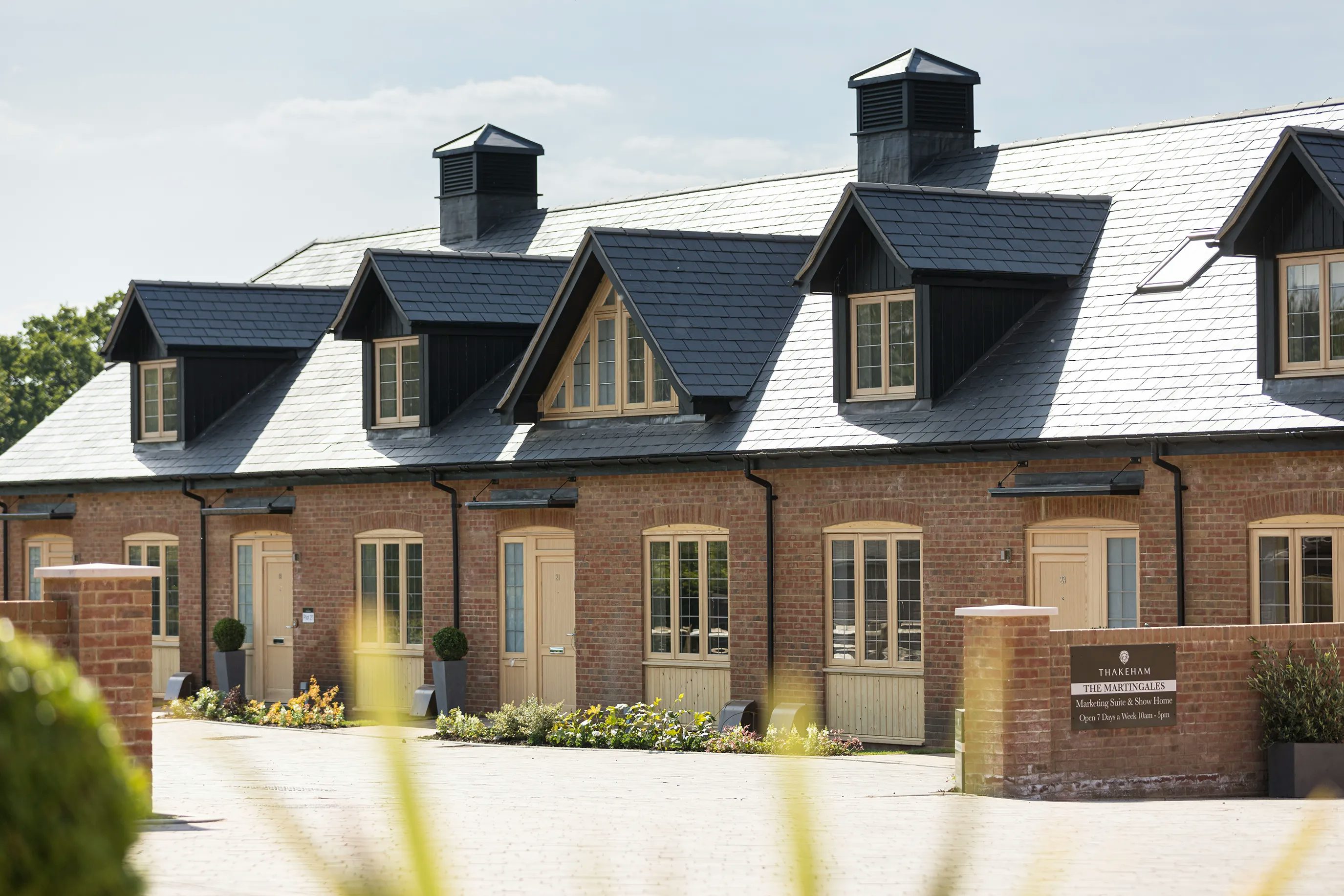 A barn-style building with a dark slate roof and wooden cladding on the exterior. Large windows and a glass entrance door are visible, and small shrubs and plants are arranged in front of the structure.