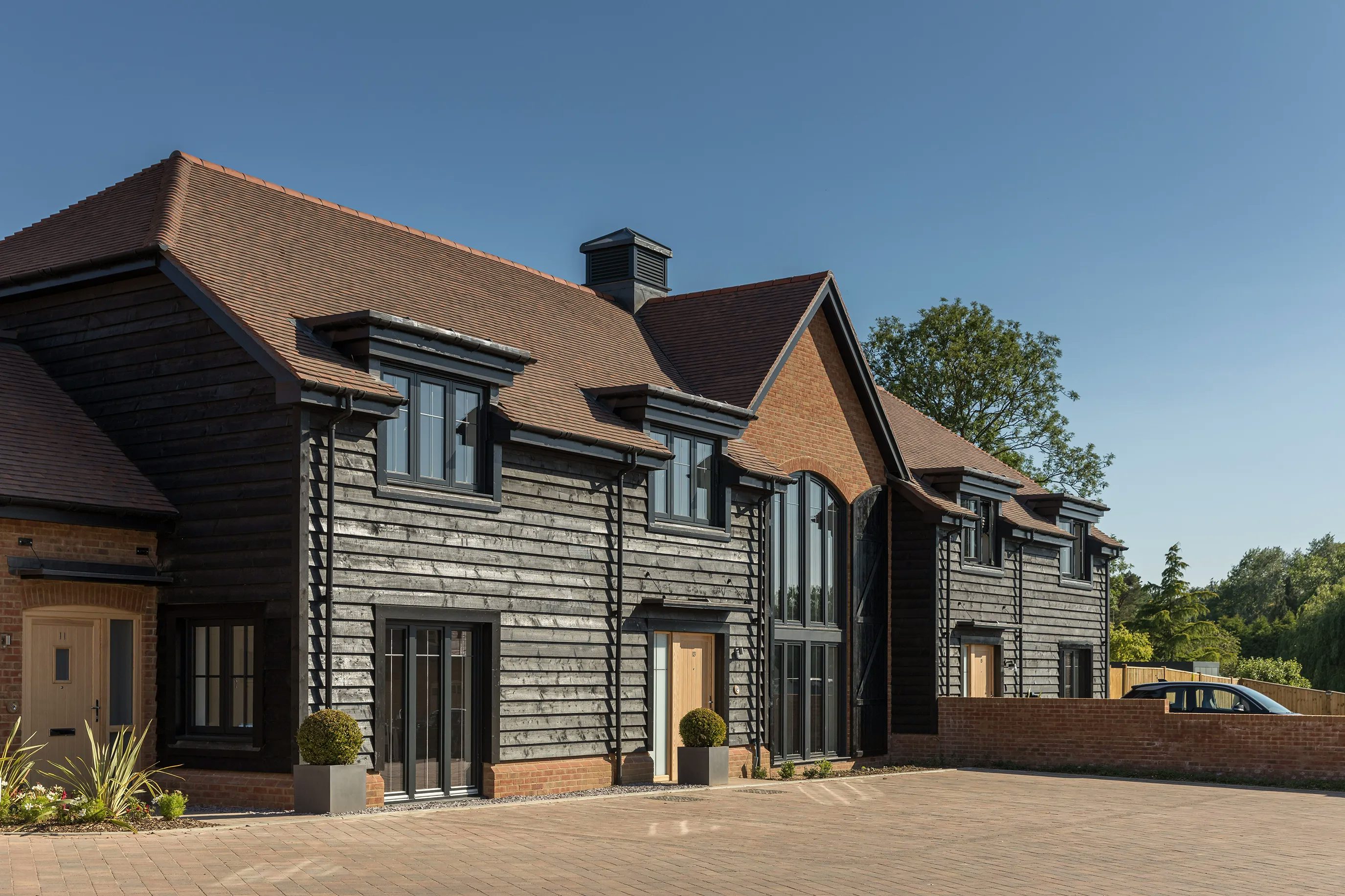 A modern residential building with a pitched red-tile roof and a combination of dark wood and brick exterior. Large glass panels in the center reflect the clear blue sky. Multiple doors and windows line the structure, with potted plants adding greenery to the paved driveway in front.