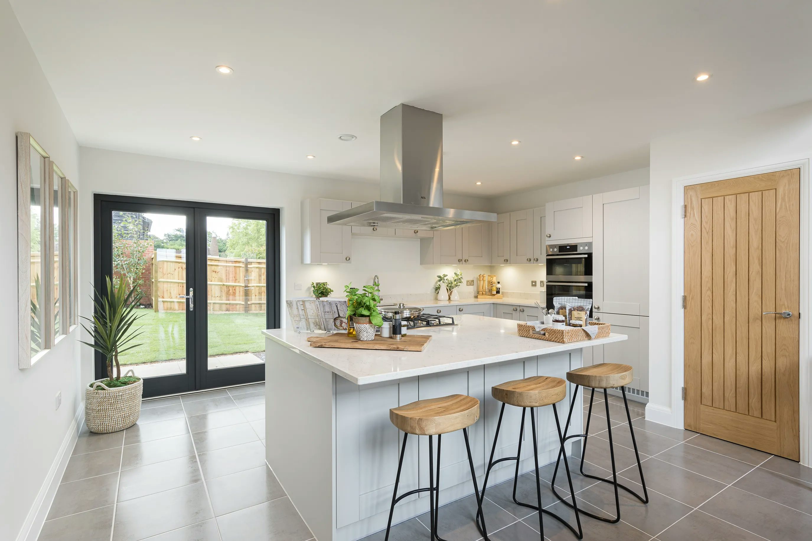 A modern kitchen with a white island in the center, featuring three wooden bar stools with black metal legs. On the island, there is a cutting board with herbs, a tray with jars, and a small plant. The kitchen has white cabinetry, a built-in oven, and a stainless steel range hood. Recessed ceiling lights illuminate the space. A wooden door is on the right, while large glass doors on the left open to an outdoor area with a lawn and wooden fence. The design is clean and contemporary.
