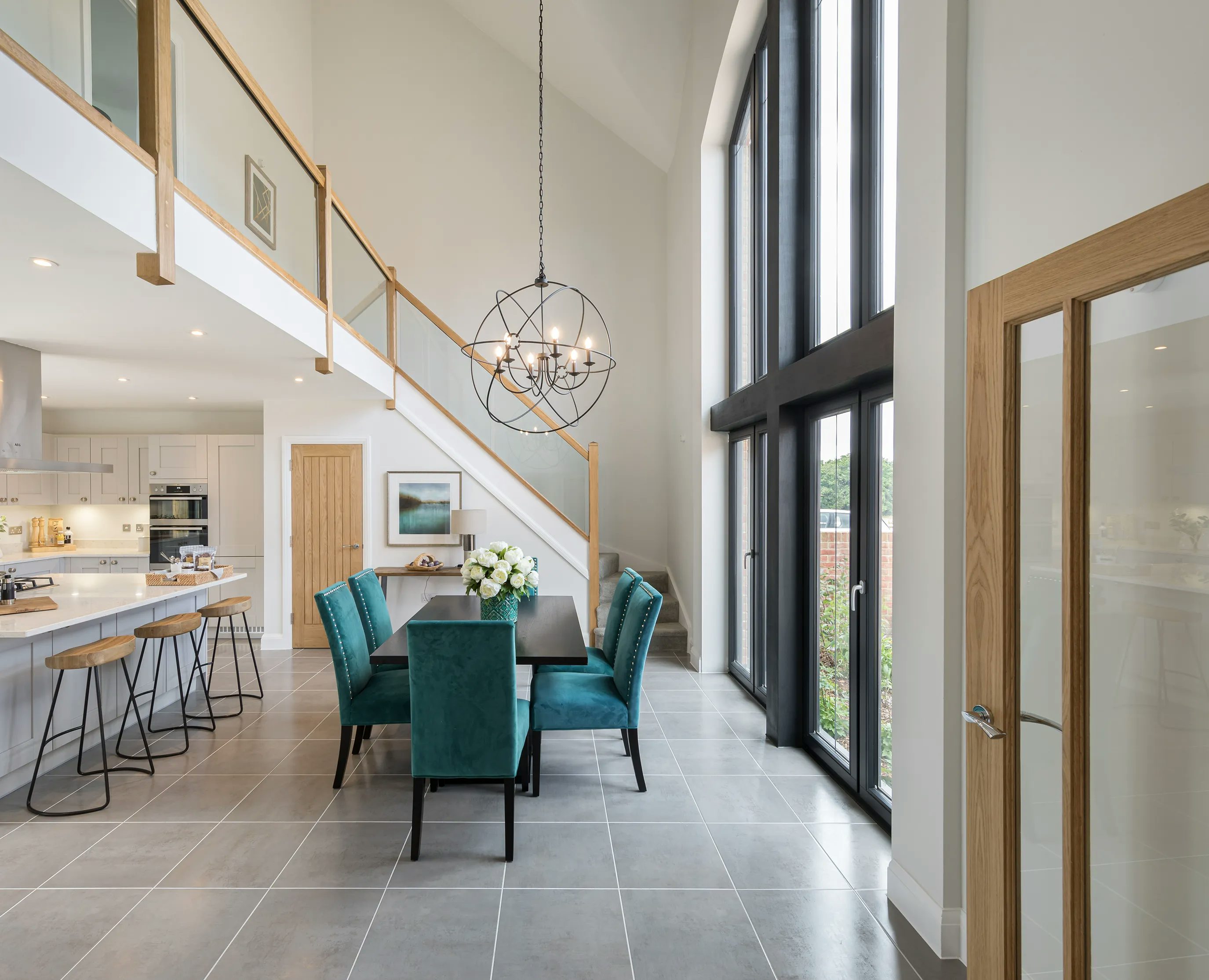 A modern, open-plan dining and kitchen area with high ceilings and large windows. The dining space features a rectangular table surrounded by teal upholstered chairs, with a white floral centerpiece. Above the table hangs a contemporary chandelier with spherical elements. The kitchen includes a long island with bar stools, sleek white cabinetry, and built-in appliances. A staircase with wooden railings leads to an upper level. Natural light streams in through large windows, creating a bright, airy atmosphere.