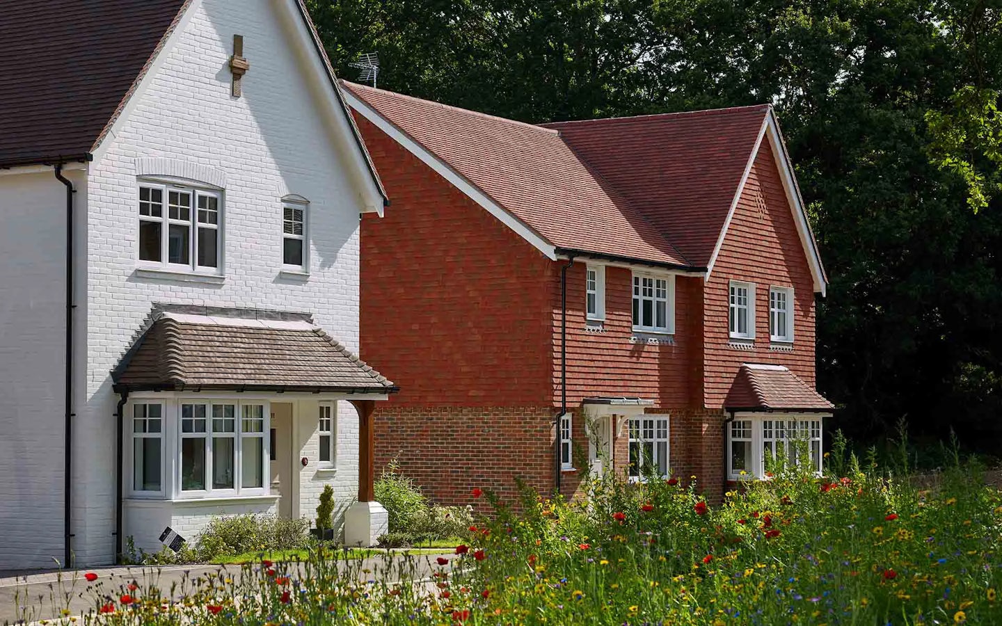 Two houses in a residential neighborhood, each with distinct architectural styles. The house on the left has white brick walls and a brown tiled roof, while the house on the right has red brick walls with a similar roof design. Both have multiple windows with white frames and small front gardens. In the foreground, vibrant flowers and greenery add color to the scene, complementing the aesthetic diversity of the homes.