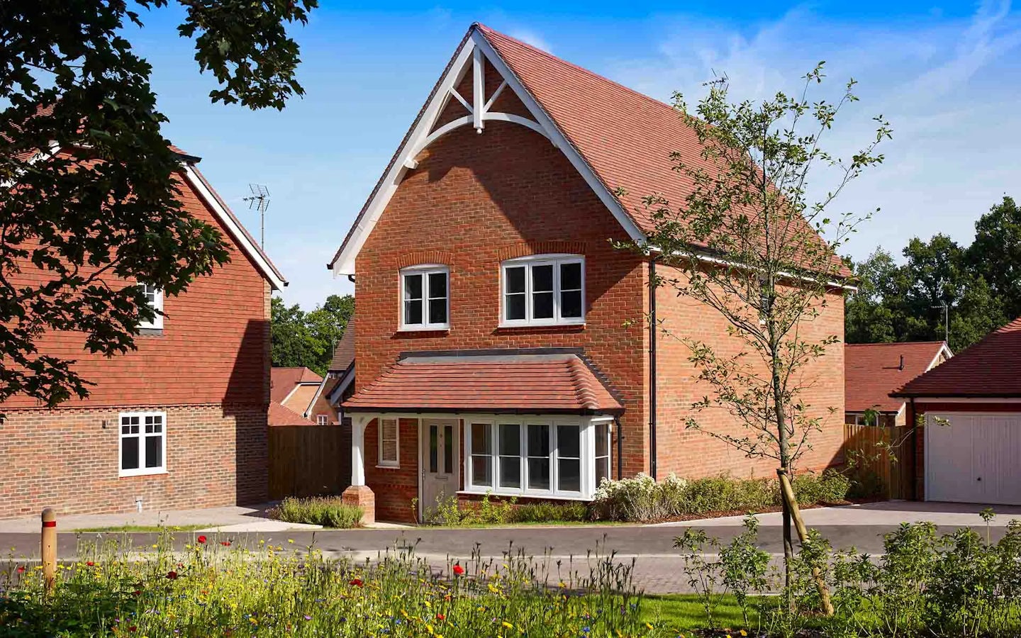 A modern, detached house with a red brick exterior and a red-tiled roof. The two-story home features three windows on the upper floor and a bay window next to the front door on the ground floor. A small garden area with a tree is near the driveway. To the left, another similar house is partially visible, and a garage is seen on the right. The sky is clear and blue, indicating a sunny day.