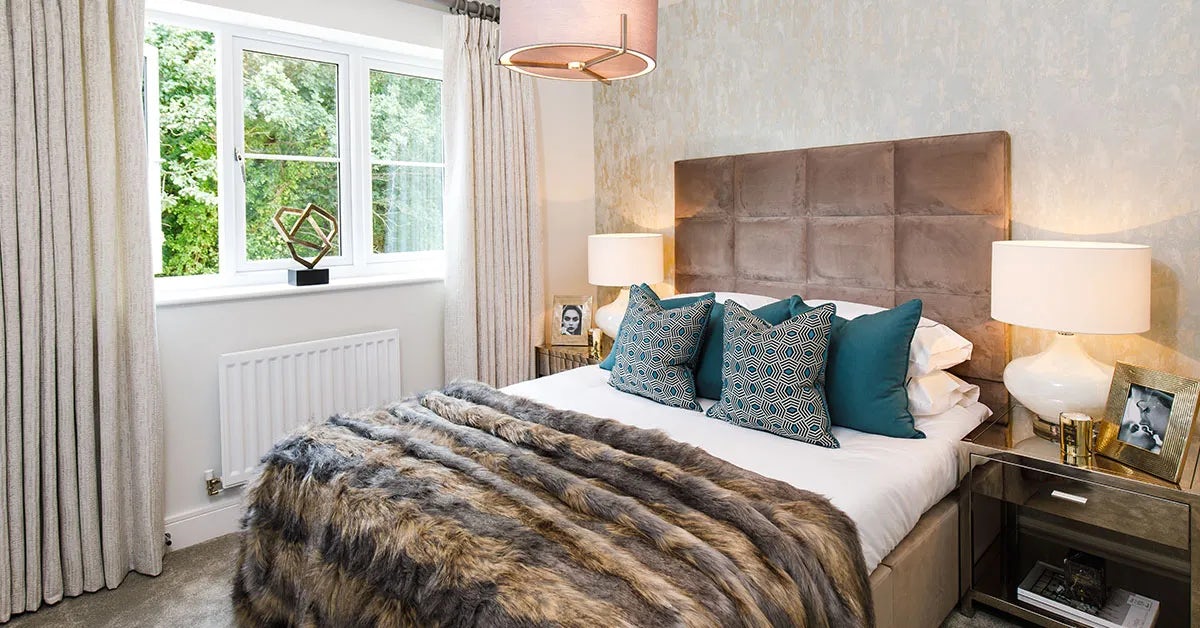 A modern bedroom with a large upholstered bed featuring teal and patterned pillows and a faux fur blanket. Two bedside tables flank the bed, each with a lamp and framed photographs. A window with beige curtains lets in natural light, revealing greenery outside. The walls have a textured finish, enhancing the elegant and sophisticated atmosphere.