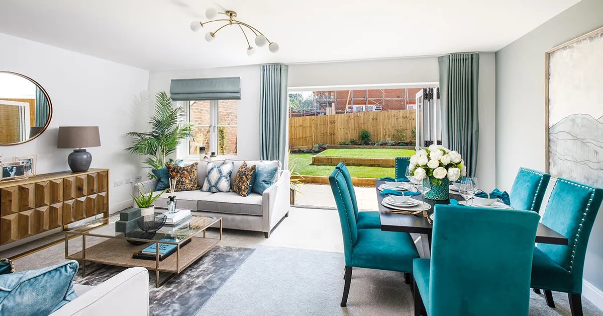 A modern living and dining area with large glass doors leading to a backyard garden. The living space features a light gray sofa adorned with decorative pillows, a glass coffee table with books and plants, and a wooden sideboard with a lamp and round mirror above it. The dining area has a dark wooden table set with plates, glasses, and a vase of white flowers, surrounded by teal upholstered chairs. Natural light fills the room through the glass doors, revealing a landscaped backyard with a wooden fence and garden beds. Light blue curtains frame the windows, and a contemporary ceiling fixture enhances the airy ambiance.